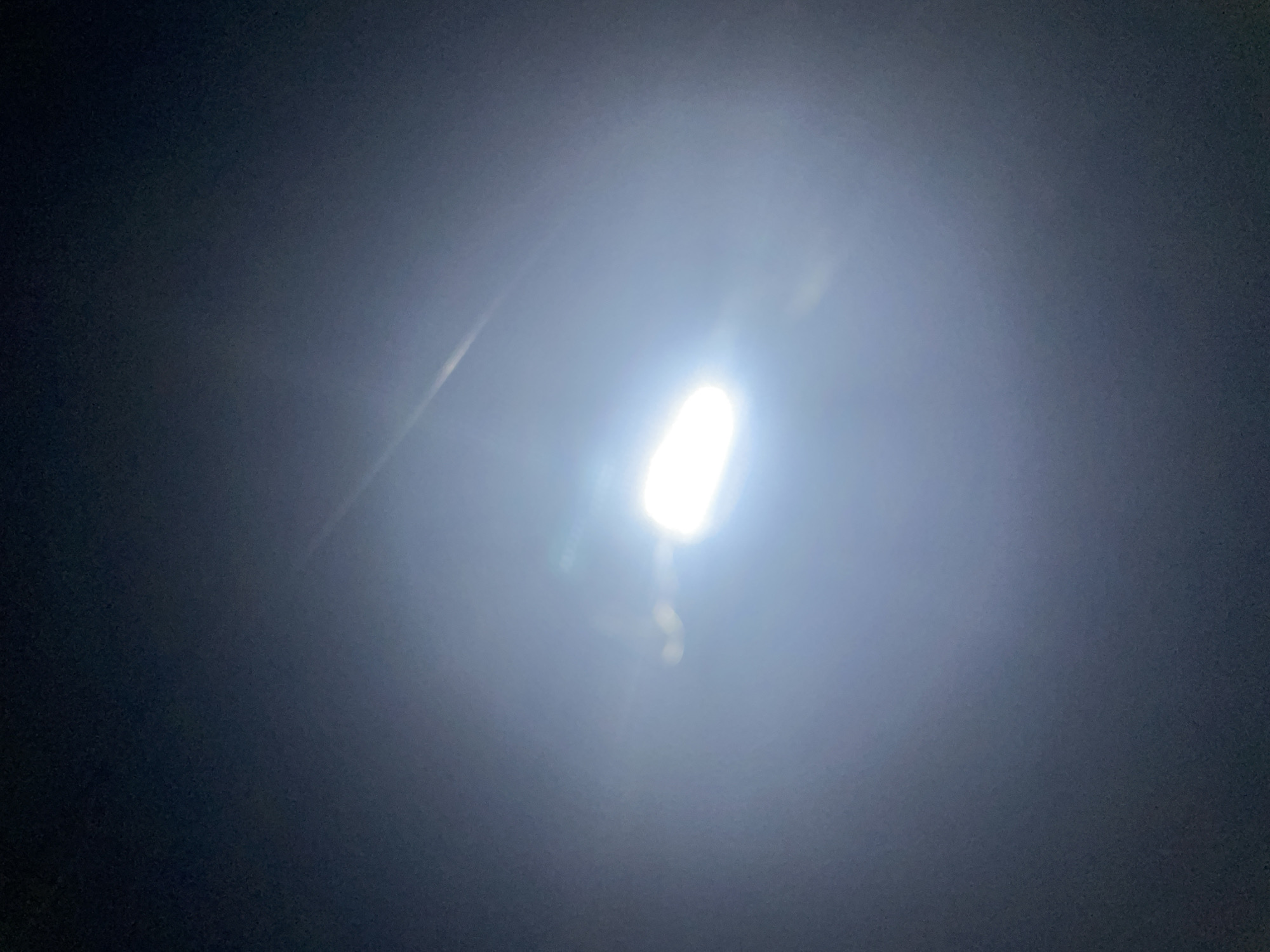 In the city of El Valle del Espíritu Santo, Venezuela, the image depicts a central, intensely bright, horizontally elongated artificial light source against an undifferentiated dark background, suggesting a nighttime environment. The light source, appearing white, emits a powerful glare that creates a prominent, expansive halo and distinct lens flares, visible as linear streaks extending diagonally upwards and to the right, and subtle circular distortions across the frame. The darkness in the surrounding areas is deep and uniform, lacking any discernible details of structures, landscape, or individuals. The composition is dominated by the radiant light and its optical effects, with the bright source positioned slightly below the vertical center.