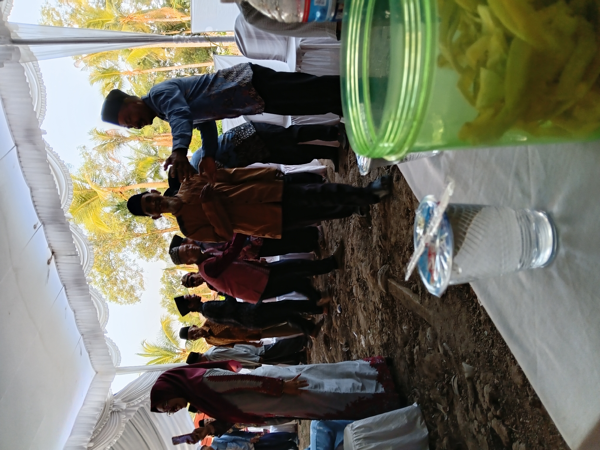 In Sumber Mulyo, Indonesia, an outdoor gathering is underway beneath a white tent structure, with palm trees and foliage visible in the background. The ground is a mix of dirt and debris. In the foreground, a clear plastic cup with a foil lid sits on a white tablecloth. To the right, a green plastic jar filled with sliced green vegetables is also on the table. A bottle of water is partially visible behind the jar. A group of men and women are present, some wearing traditional Islamic attire including peci hats. One woman in the foreground wears a maroon hijab and a light-colored dress. Several men are interacting, with one in a blue patterned shirt shaking hands with another in a tan shirt. The overall scene suggests a social event or celebration.