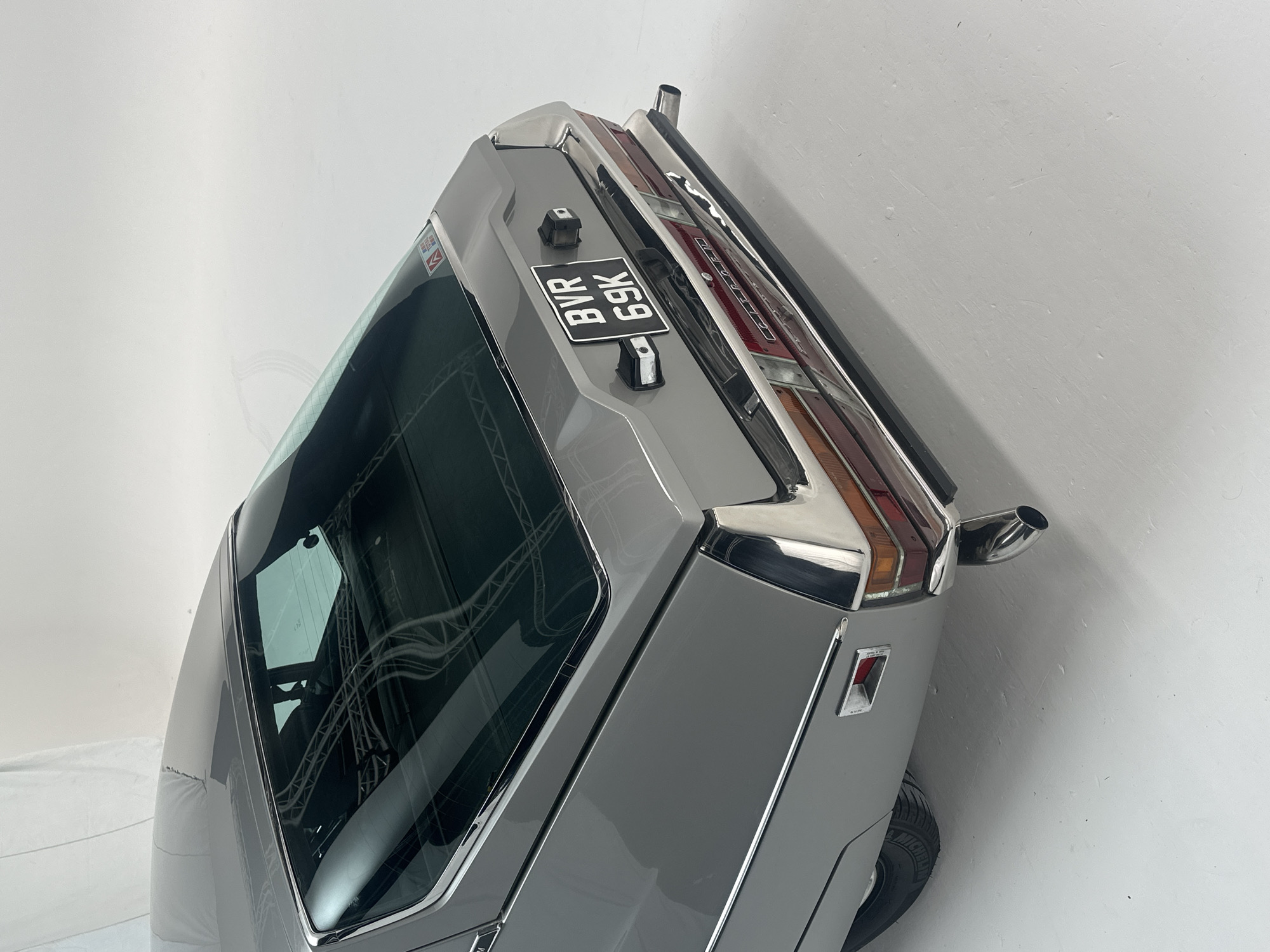 The rear section of a vintage grey car is depicted at an angle, positioned against a plain white background, suggesting an indoor studio or exhibition setting in London, United Kingdom. The vehicle is stationary, presented for display.
The car features a prominent chrome bumper and trim that frames the tail lights and extends along the rear, highlighting its classic design. The tail lights themselves are multi-sectioned, featuring red and amber lenses. A single, polished chrome exhaust pipe exits from the right side of the rear.
A black UK license plate, clearly displaying "BVR 69K" in white characters, is mounted centrally on the trunk lid, flanked by two black rubber or plastic buffers. The rear window, slightly tinted, reflects a complex structure of metal trussing or scaffolding, further indicating an artificial, controlled environment. A small, partially obscured sticker with a red "M" logo is visible on the upper left corner of the rear window. A portion of a tire, possibly a Michelin given the faintly visible text on the sidewall, is visible on the lower right, contributing to the car's grounded appearance. The car's paintwork appears well-maintained, reflecting light evenly across its surfaces.