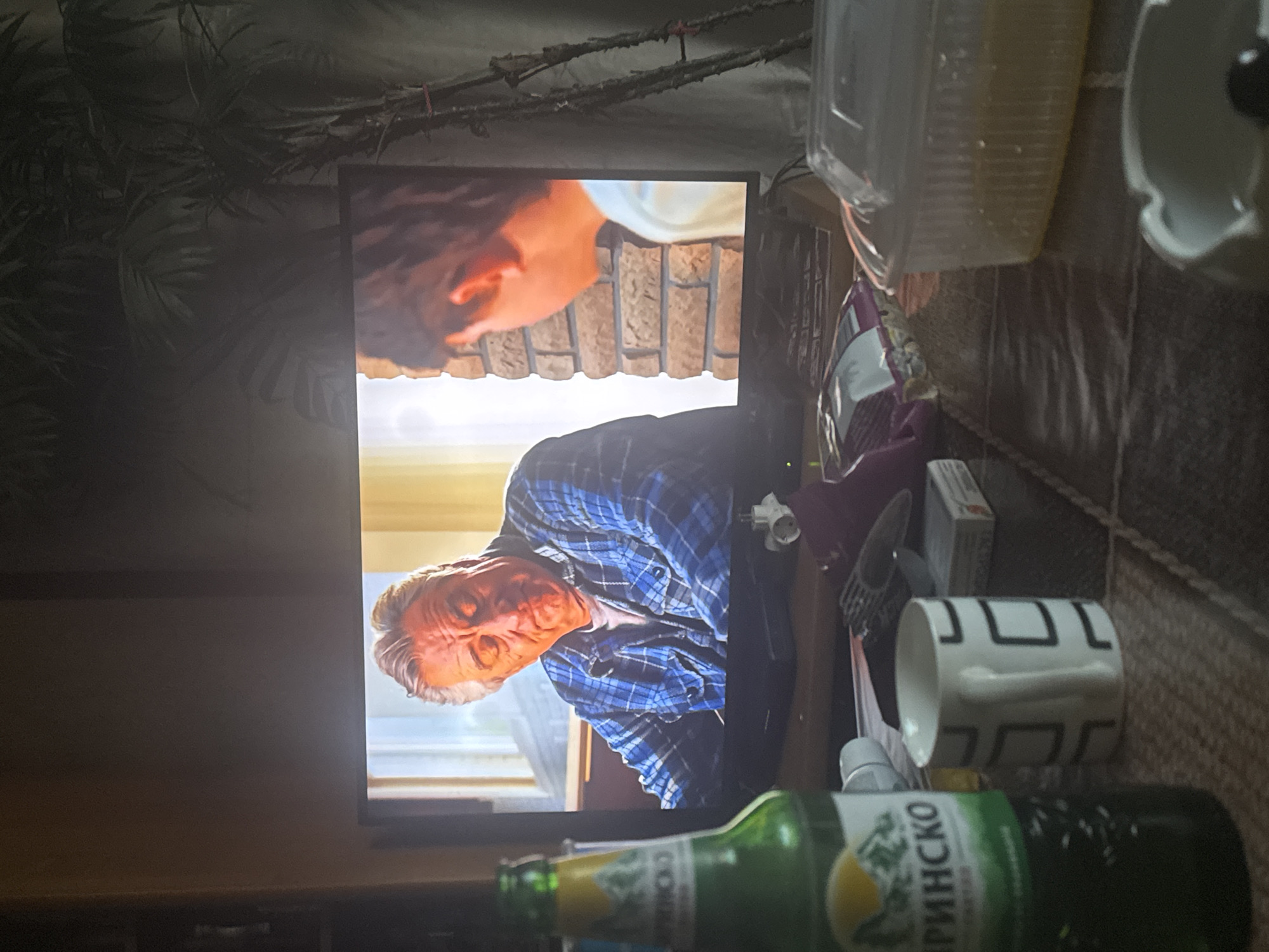 A television screen displays a scene from a film, featuring two individuals. In the foreground, on a table, are several objects: a green glass bottle with a label depicting a mountain landscape, a white mug with a geometric pattern, a packet of cigarettes, a clear plastic container with food, and an ashtray. Behind the television, a large artificial palm plant is visible. The scene appears to be indoors, likely a living room, given the presence of the television and furniture. The overall lighting suggests it is evening or nighttime, with the television screen being the primary light source. The room's ambiance suggests a casual, domestic setting.