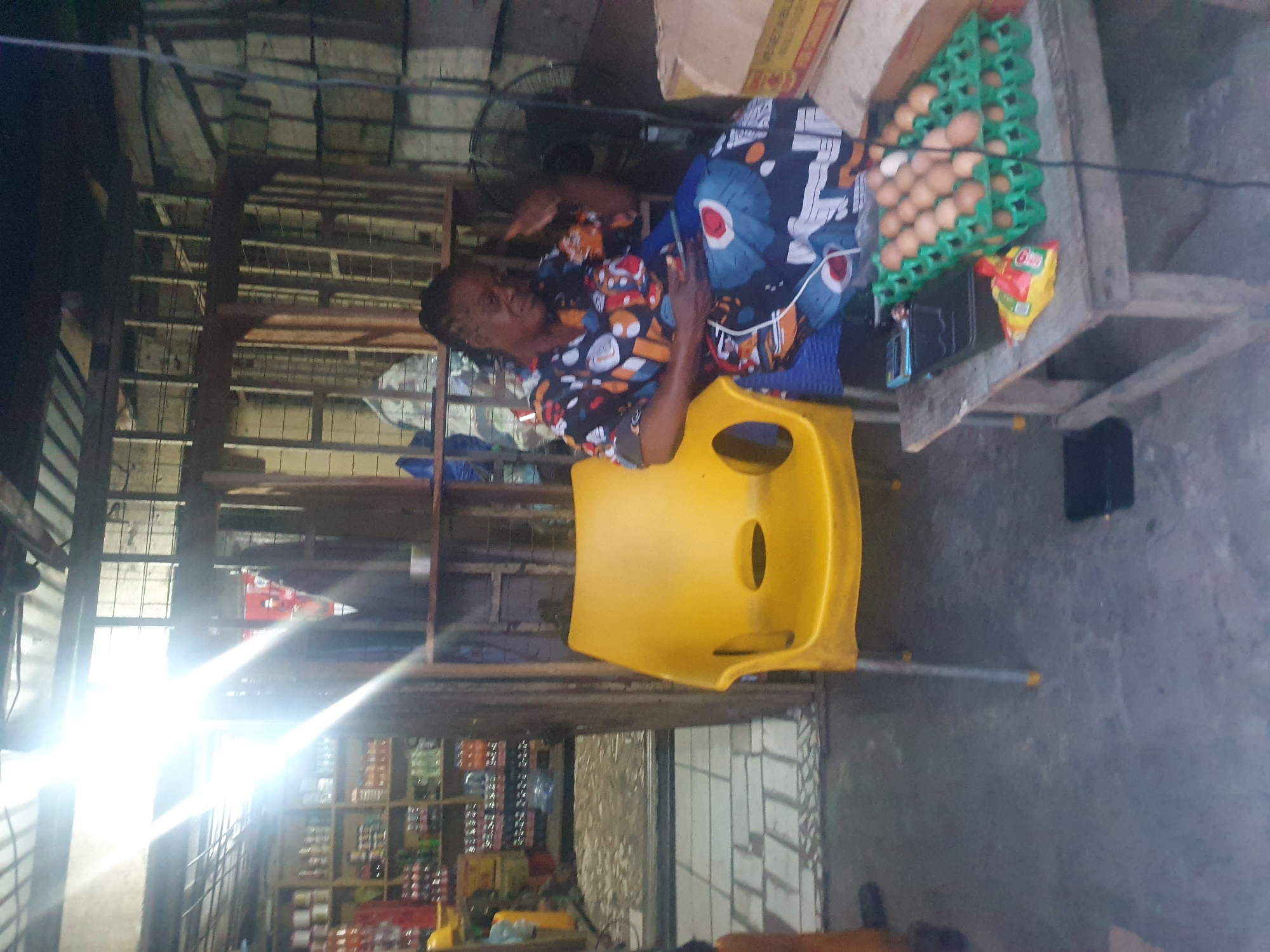 An elderly woman sits on a yellow plastic chair in what appears to be a small shop or stall. The shop is filled with goods on shelves. To her right, there is a table holding a carton of eggs and a snack. The exact location within Lokoja, Nigeria, cannot be determined.