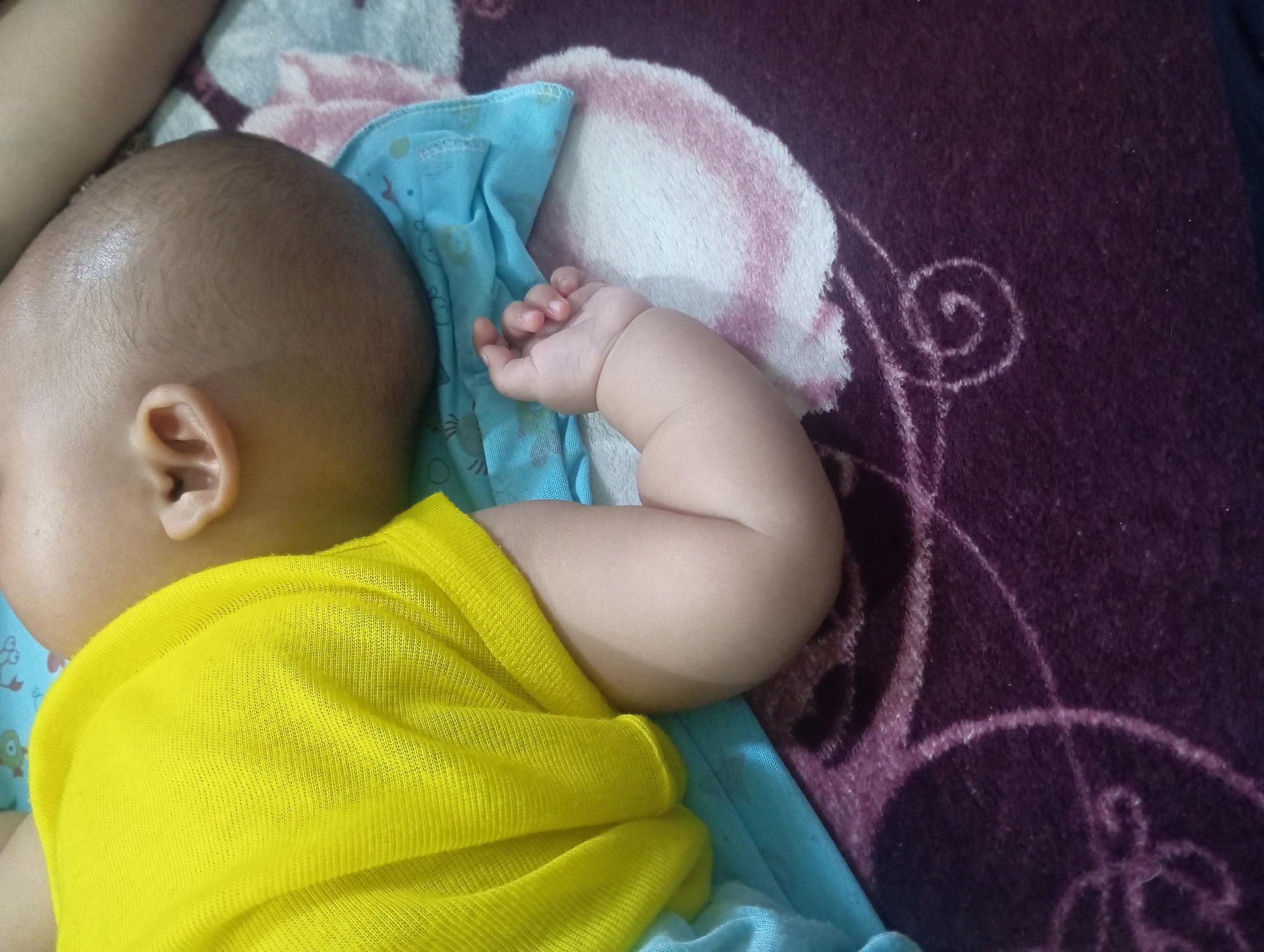 The image shows a close-up view of a baby sleeping on its side. The baby is wearing a yellow t-shirt. The baby's arm is bent and its hand is resting on a light blue cloth. The baby is lying on a patterned rug. The background is out of focus. The image appears to have been taken indoors, possibly in a home in Rejosari, Indonesia.