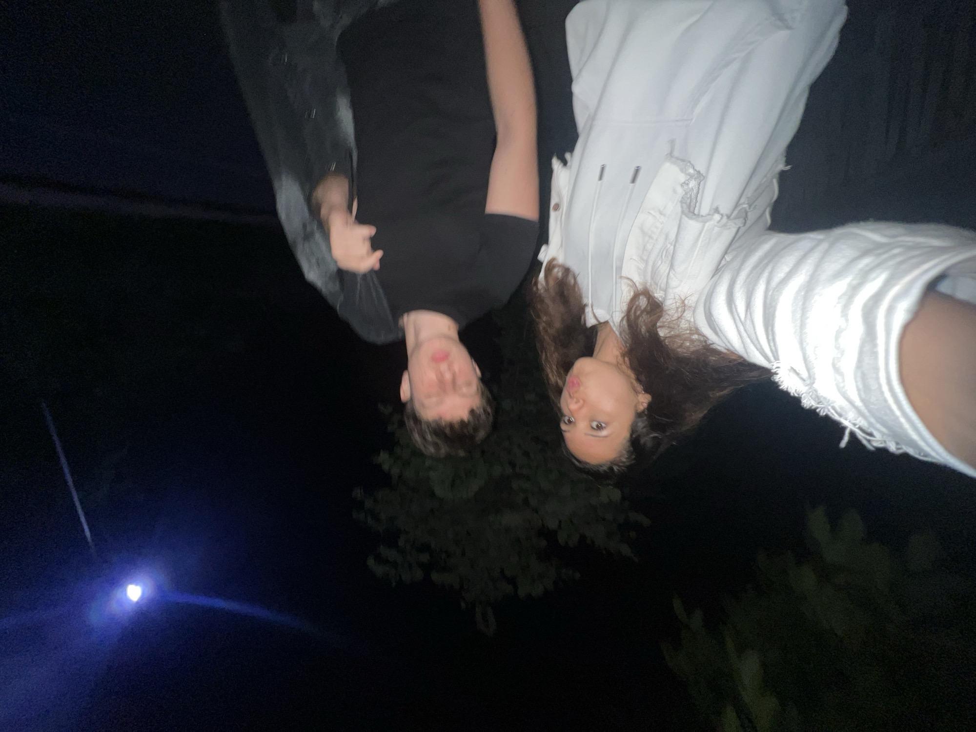The image captures two individuals, a female and a male, outdoors at night in an unknown city and location. The female, positioned closer to the camera on the left, has long, dark, wavy hair and is wearing a white hooded sweatshirt under a distressed white denim jacket. She is taking a selfie, with her lips pursed in a "kiss" pose and her eyes looking slightly upwards. To her right, the male stands slightly behind her, wearing a black short-sleeved t-shirt. His eyes are closed or heavily squinted, and he holds a dark jacket draped over his right shoulder and arm. The background is dark, with the silhouette of trees visible. In the upper right quadrant, a bright, blueish-white street lamp on a pole emits a noticeable lens flare.