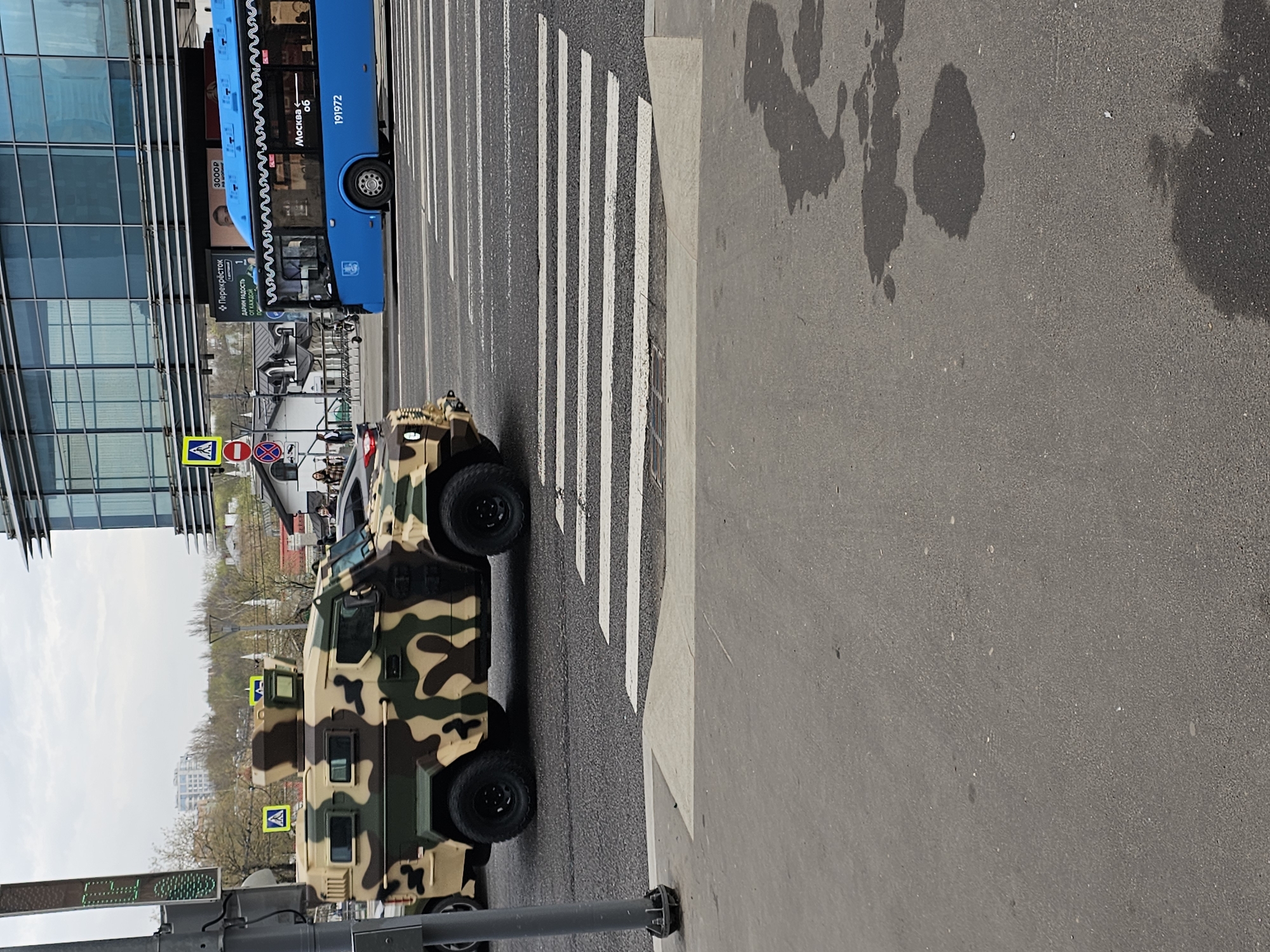 The image captures an urban scene, likely in Khimki, Russia, given the presence of a blue Moscow bus partially visible on the left side of the frame. The bus is parked next to a modern glass-walled building. A camouflage-painted armored vehicle is positioned in the middle of a pedestrian crosswalk, also known as a zebra crossing. The ground is asphalt with visible water stains and marks. The sky is overcast. A pedestrian crossing sign is visible in the background, along with a traffic light pole. No people are distinctly visible in the image.