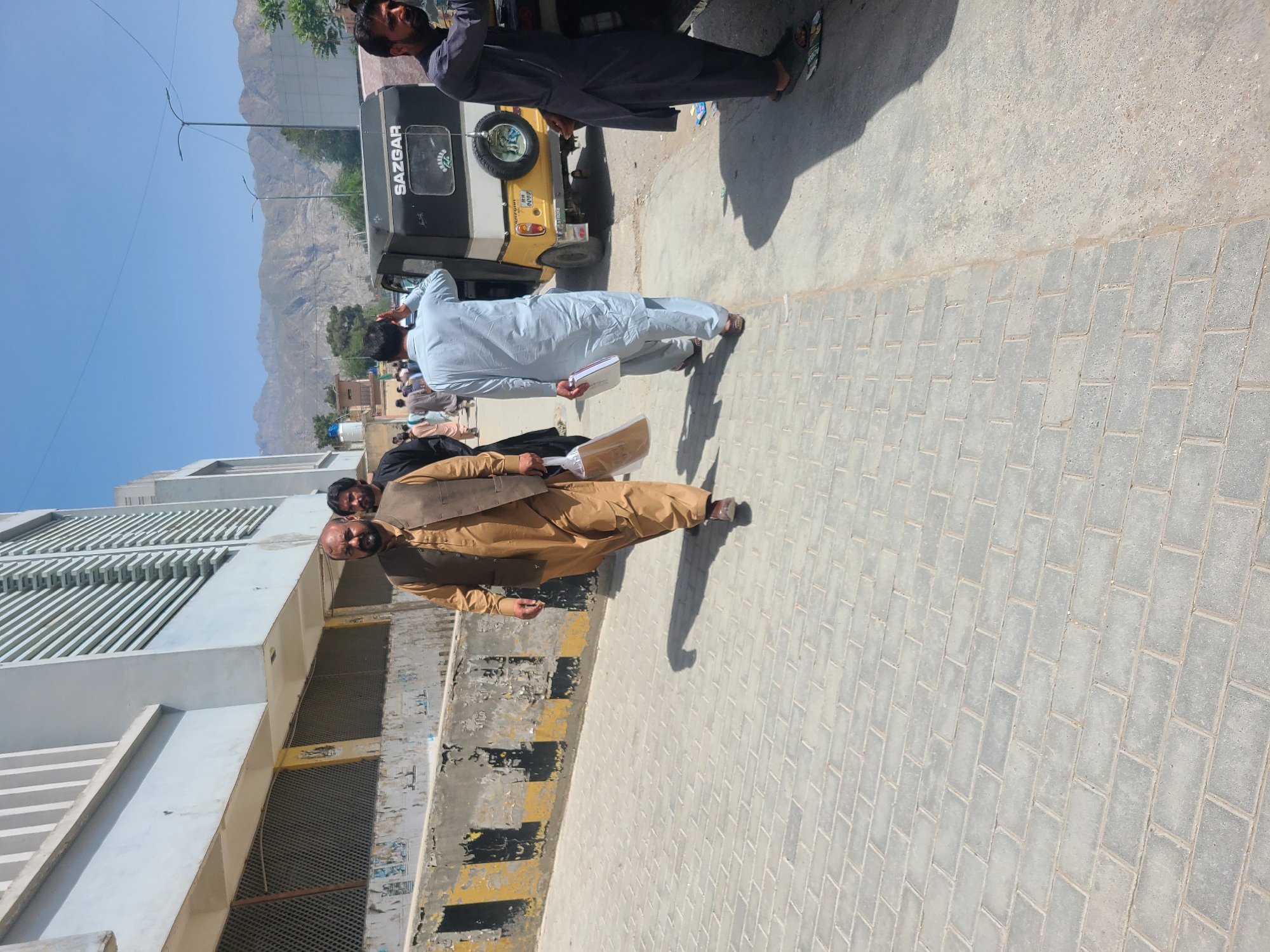 The image shows a street scene in Quetta City Tehsil, Pakistan. Several men are present, some wearing traditional clothing. A yellow and black "SAZGAR" three-wheeled vehicle is visible in the background, along with mountains in the distance. A building with a modern facade stands to the left, displaying weathered paint and peeling sections. The ground is paved with interlocking blocks.