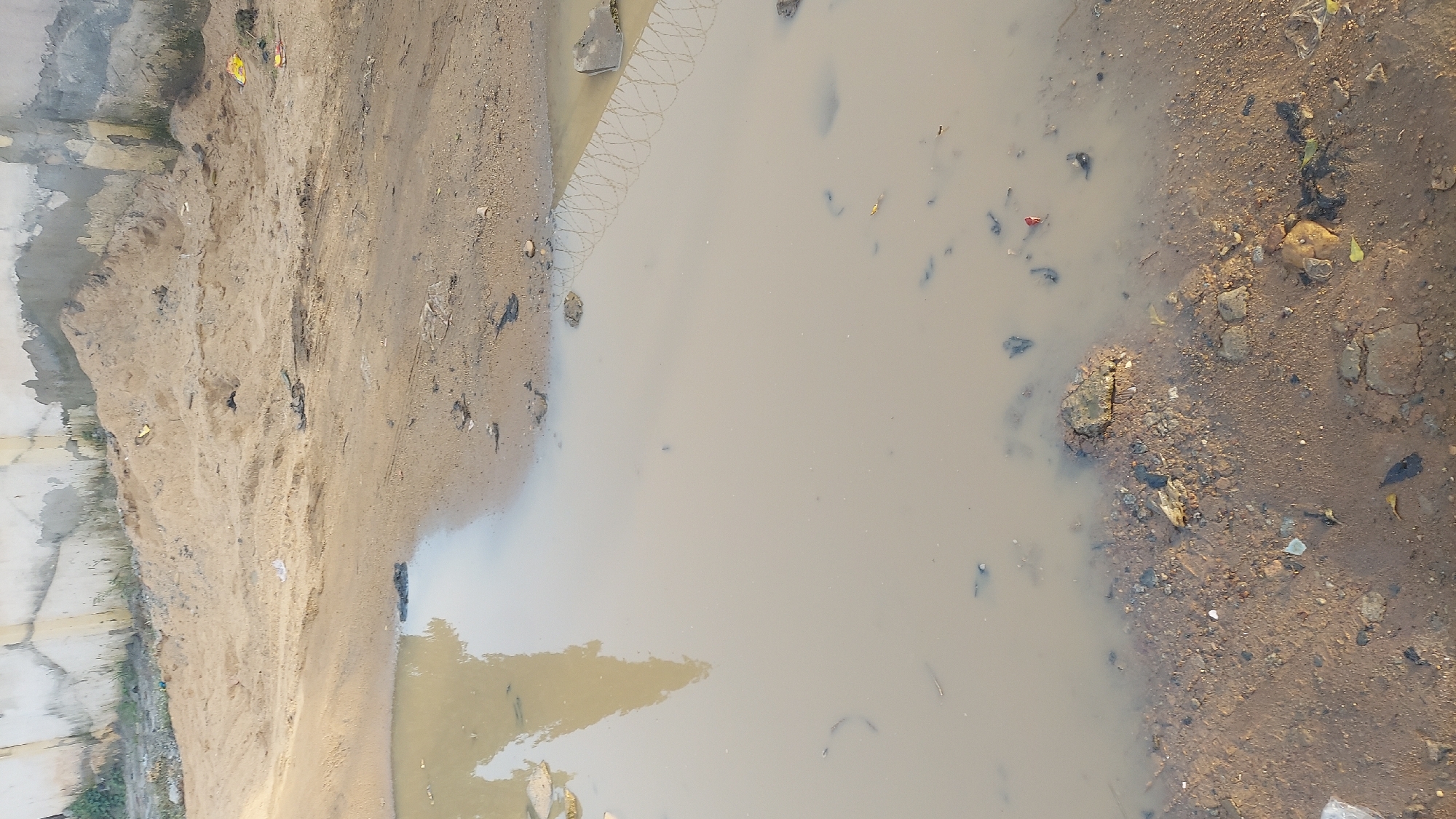 The image depicts an outdoor scene in Abuja, Nigeria, featuring a large, murky puddle dominating the foreground and midground. The water is a light brown color, suggesting it is mixed with sediment and possibly runoff. The edges of the puddle reveal a rough, uneven ground composed of dirt, gravel, and scattered debris, including small rocks and fragments of what appear to be plastic.
In the background, a sandy embankment rises, covered with scattered litter such as plastic bags and wrappers, indicative of an unmanaged or undeveloped area. Above the embankment, a section of a concrete wall is visible, showing signs of wear and tear, with some patches of discoloration and possibly algae growth. The overall impression is one of a neglected outdoor space, likely a roadside or undeveloped plot, with the puddle being a consequence of recent rainfall or poor drainage. There are no people or active events captured in the image.