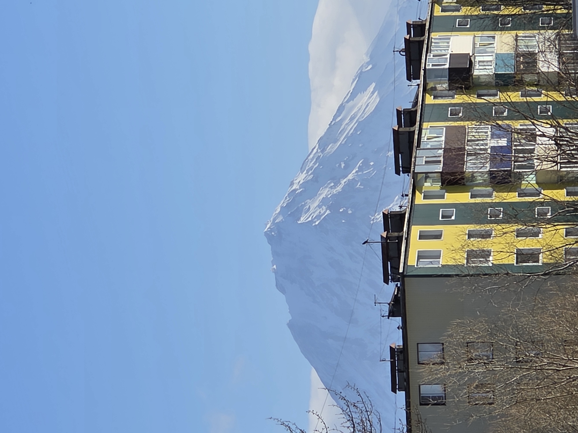 The image shows a high-rise apartment building with yellow and green paint. A large snow-capped mountain is in the background, partially obscured by clouds. The sky is blue. The trees in the foreground are bare. The location is Unknown, Unknown.