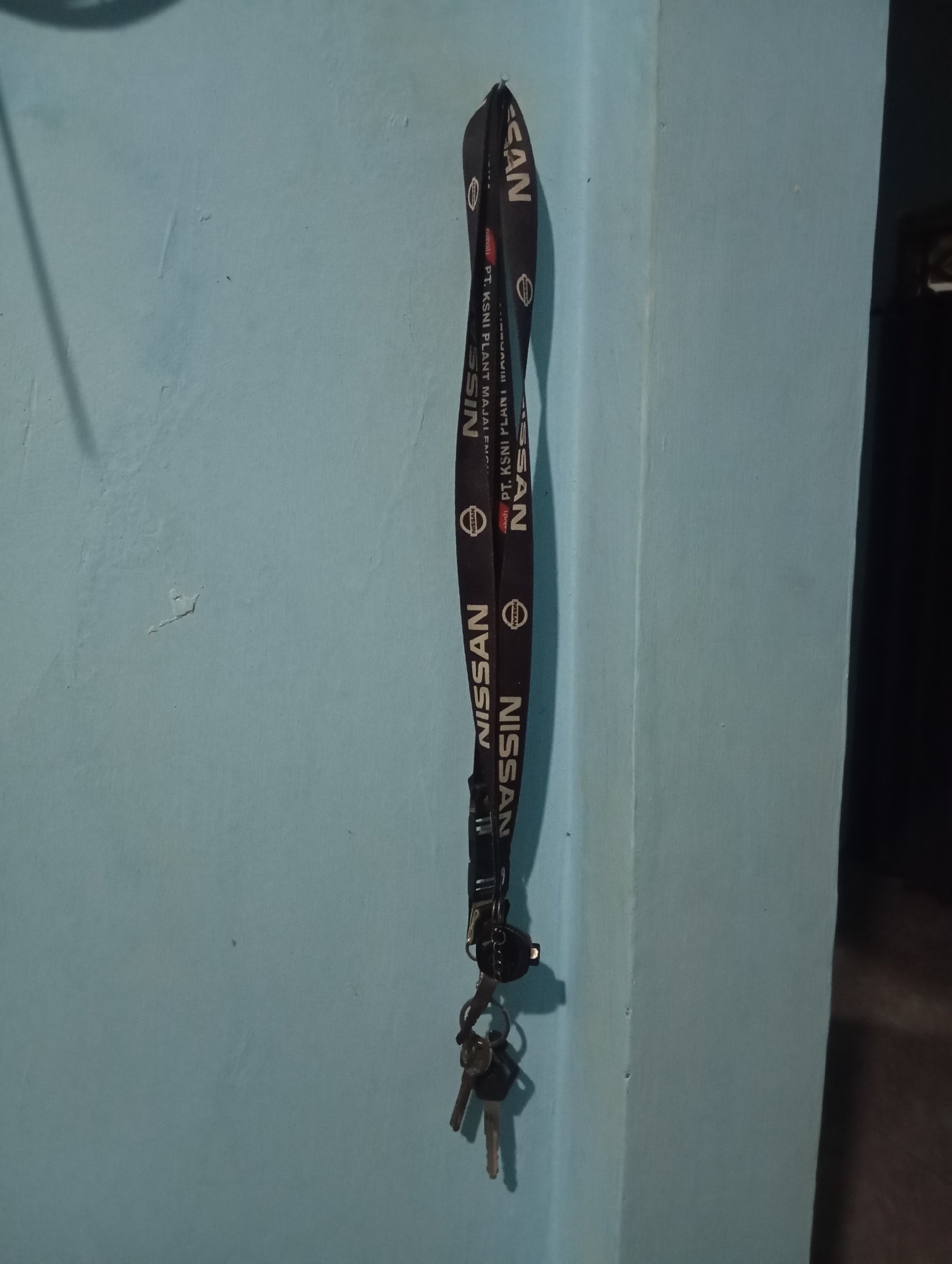 A close-up, vertical, eye-level shot captures a lanyard with keys hanging on a light blue wall. The lanyard, which appears to be black, is adorned with the "NISSAN" logo and text. At the top of the lanyard, "CAN" is visible, and below it, "PT. KSNI PLANT MAJALENGKA" can be read. Further down, the "NISSAN" logo appears multiple times. At the bottom of the lanyard, a set of keys is attached. The wall itself shows some subtle texture and imperfections. The lighting in the scene is dim, with shadows cast by the lanyard and keys onto the wall. The background on the right is dark, suggesting an interior space or the edge of a room. The image is set in Sudikampiran, Indonesia.