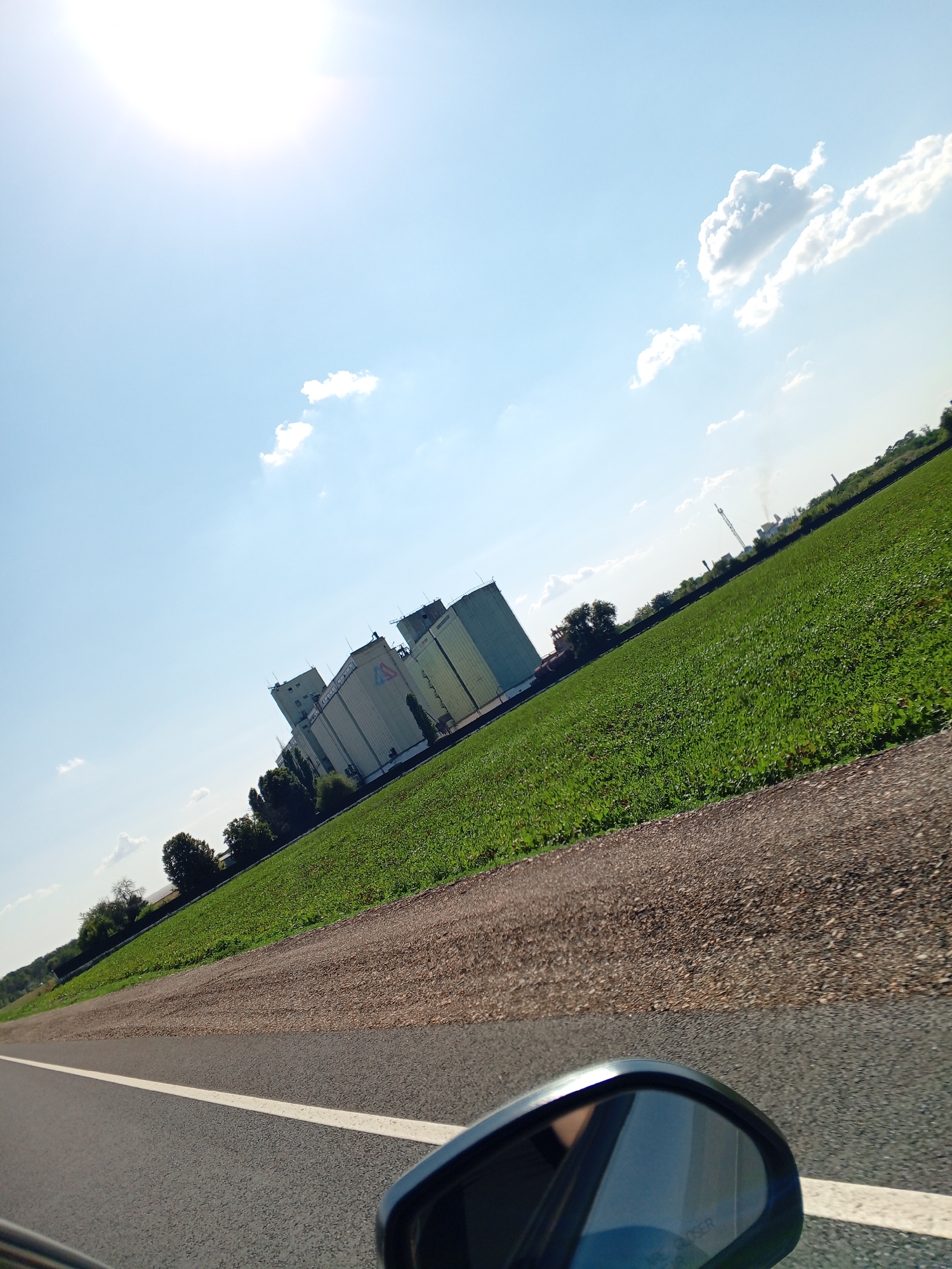 The image, taken from a vehicle's side mirror, shows a tilted view of a rural or industrial landscape under a bright, sunny sky with scattered clouds. The foreground features a segment of an asphalt road with a white line, bordered by a gravel shoulder. To the right of the road is a vast expanse of green farmland, appearing as a field of crops. Beyond the field, a row of large, light-colored industrial silos or storage buildings is visible, some featuring visible logos or markings. The structures are tall and rectangular, suggesting an agricultural or processing facility. Further in the distance, trees and other industrial elements, possibly including a crane and smokestacks, dot the horizon. The overall scene conveys a sense of open space and activity, likely a roadside view of an industrial agricultural area.