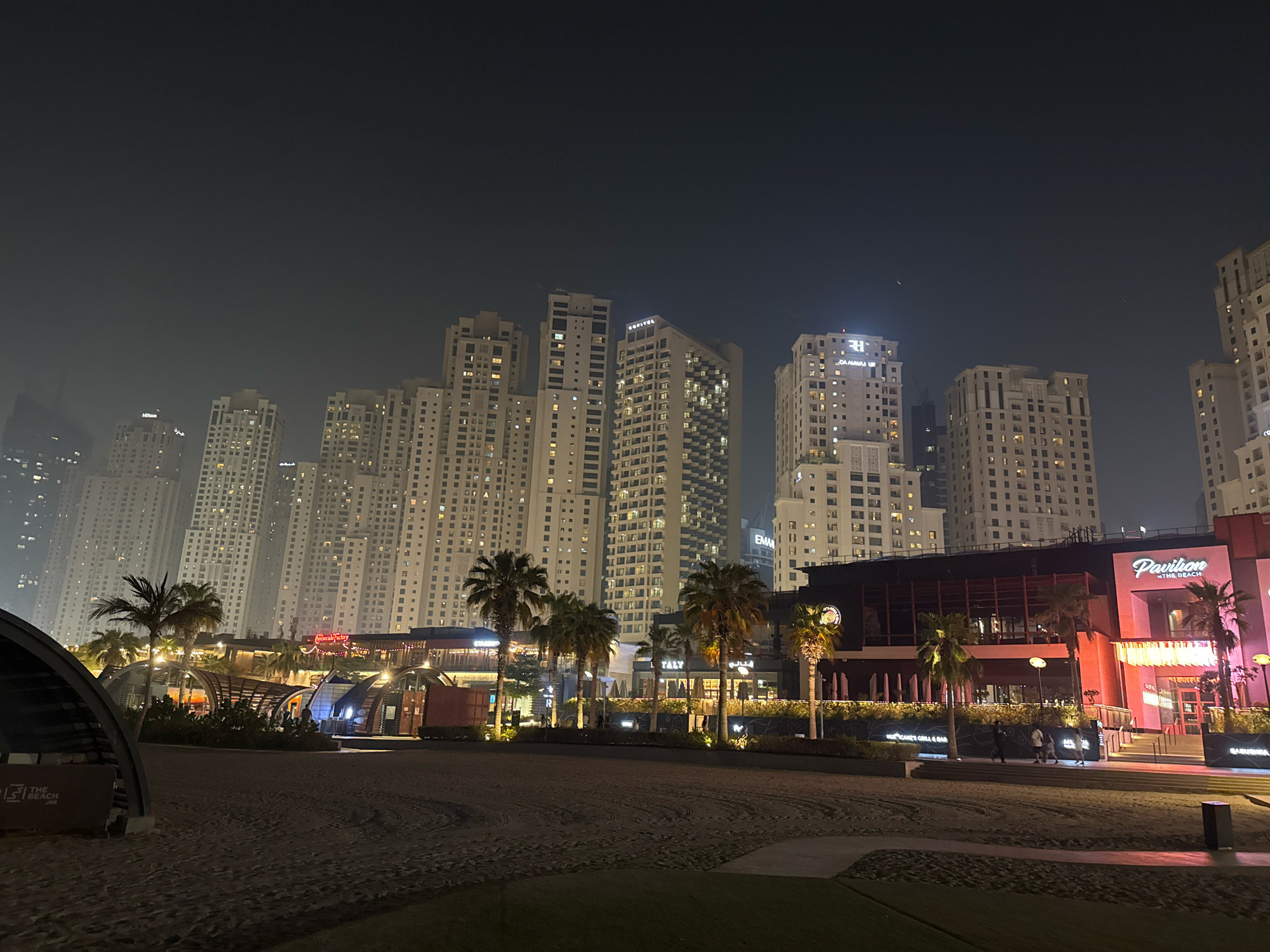 This is a night photograph of the Jumeirah Beach Residence (JBR) area in Dubai, United Arab Emirates. The scene is dominated by a dense cluster of modern, high-rise residential buildings illuminated by countless windows. In the foreground, a sandy beach is visible, with tire tracks imprinted on its surface, suggesting recent vehicular activity or maintenance.
To the left of the frame, a large, dark, curved structure, possibly an architectural element or part of a beachside venue, partially obstructs the view. Further to the right, a strip of low-rise buildings housing restaurants and shops lines the waterfront. Notable signage includes "Pavilion @ THE BEACH" in a prominent pink building, and other establishments like "Cheesecake Factory" and what appears to be "Starbucks". Palm trees are scattered throughout the area, their silhouettes stark against the illuminated buildings. Several people are visible walking along the promenade in front of the restaurants. The overall atmosphere is one of urban nightlife and leisure, with the imposing skyscrapers of the Dubai Marina skyline serving as a backdrop. The sky is dark, with a slight haze obscuring the upper portions of the tallest buildings.