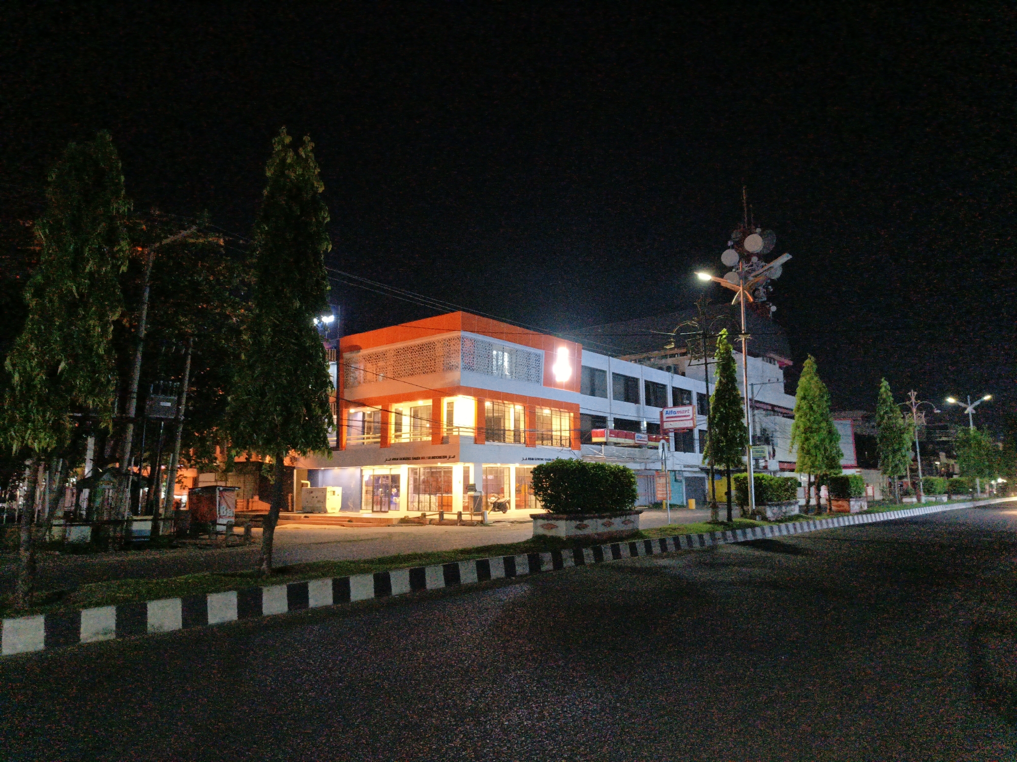 The image shows a nighttime street scene in Lhokseumawe, Indonesia. The focus is on a two-story building, lit with orange and white lights. The building has an orange top and a white lower section. A lighted symbol is visible on the building. Trees line the street on both sides. A road with black and white striped curbs is in the foreground. Overhead streetlights illuminate the scene. A telecommunications tower is visible near the building. An "Alfamart" sign is visible on a building adjacent to the lighted building. The night sky is dark.