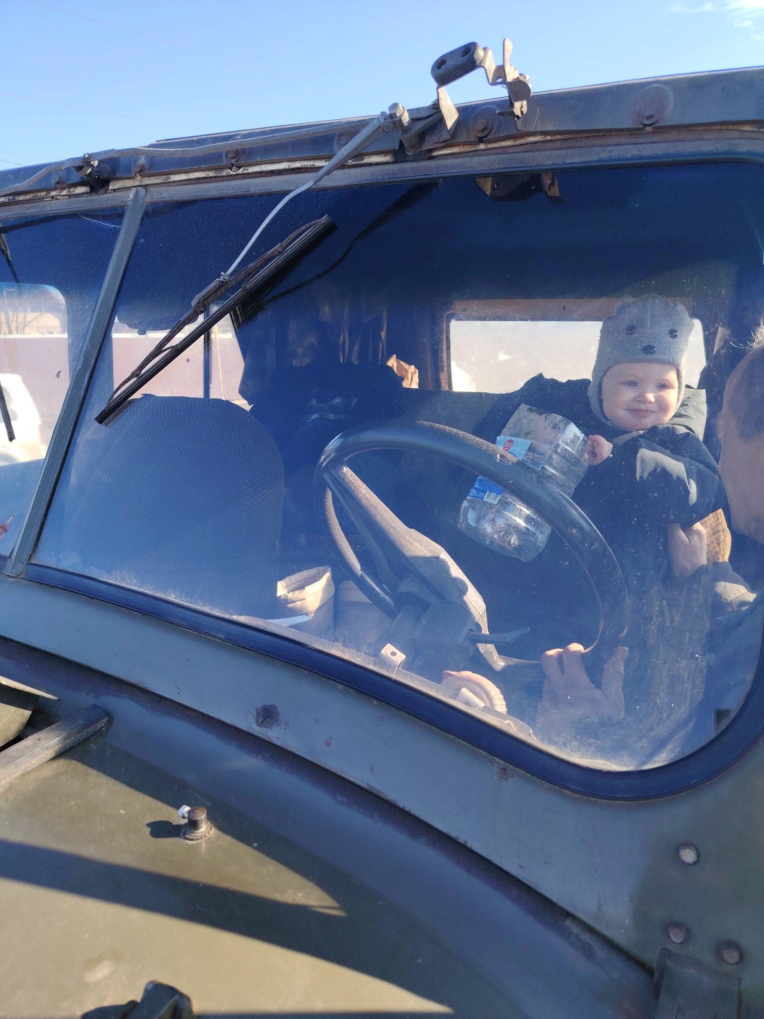 A child is sitting at the steering wheel of a military vehicle. The child wears a grey knit hat with bear ears, and a dark jacket. There is an adult next to the child, holding them. Two plastic water bottles sit on the steering wheel. The vehicle is olive green, with a dirty windshield, and is likely located in Chaikovsky, Russia.