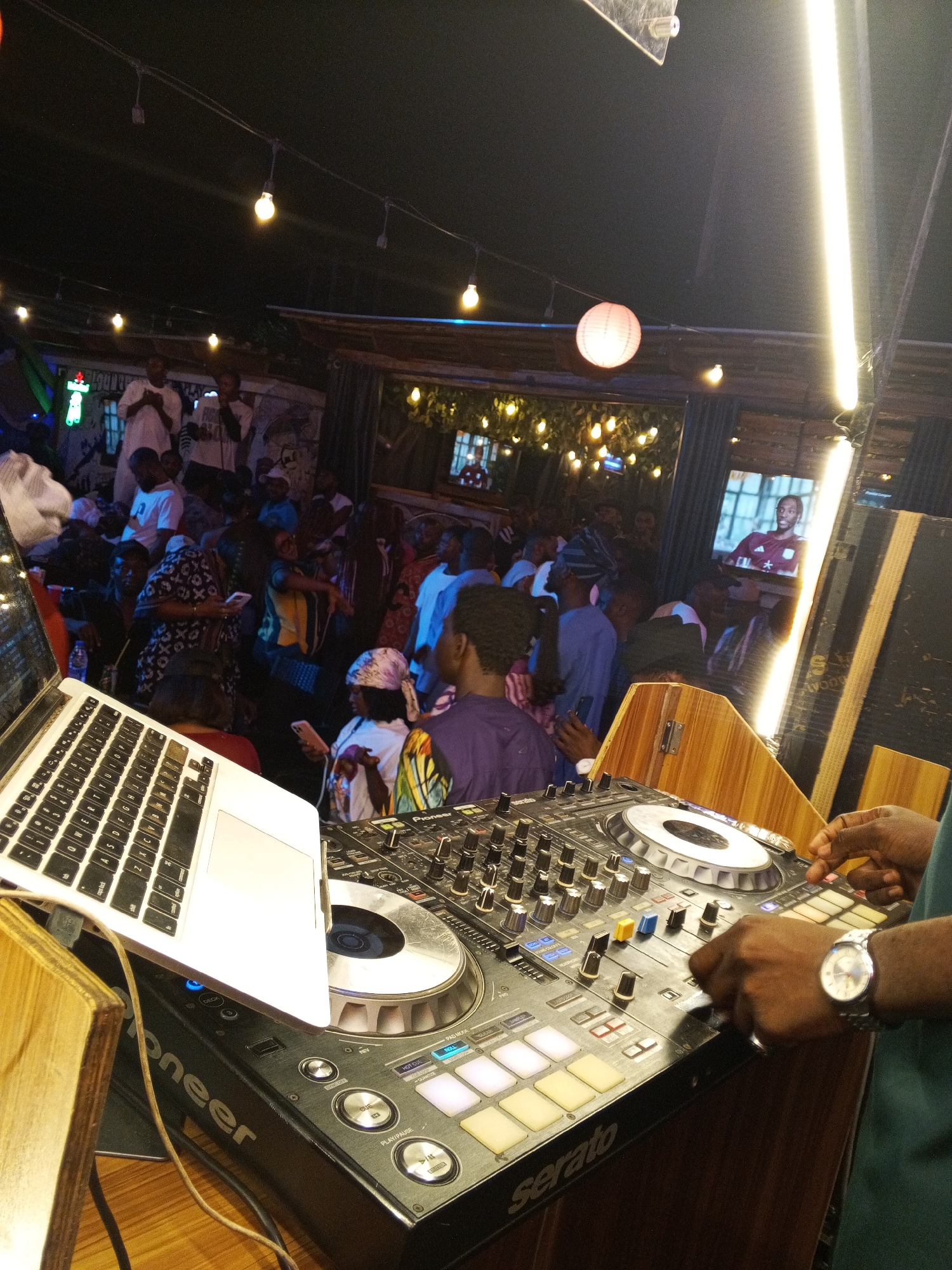 Here is a detailed description of the image:
**Scene:** The image appears to be taken inside a busy nightclub or party venue. A DJ booth is prominent in the foreground, with a laptop and DJ mixer. Behind the DJ setup, a crowd of people is visible, suggesting a lively atmosphere with music and dancing.
**People:** A DJ is operating the equipment. In the background, there are many people, most of whom appear to be dancing or socializing. The people in the crowd seem to be of various ages and wearing different styles of clothing.
**Objects:** The main objects include a laptop, a professional DJ mixer (labeled "Pioneer" and "Serato"), wooden panels (possibly part of the DJ booth), lights, a round paper lantern, and screens showing video content.
**Actions/Interactions:** The DJ is actively mixing music, indicated by the hands on the mixer. The people in the background are dancing, talking, and generally enjoying the music.
**Environment:** The venue has a nightclub or bar-like appearance, with decorative lighting and screens. The crowd suggests that it is a popular spot for socializing and dancing. The lighting is a combination of hanging bulbs and a bright vertical light on the right, creating a lively ambiance.
**Location:** The location in the city of Unknown, Unknown is not explicitly stated. However, based on the club-like setting and the DJ equipment, it could be in an entertainment district of the city.