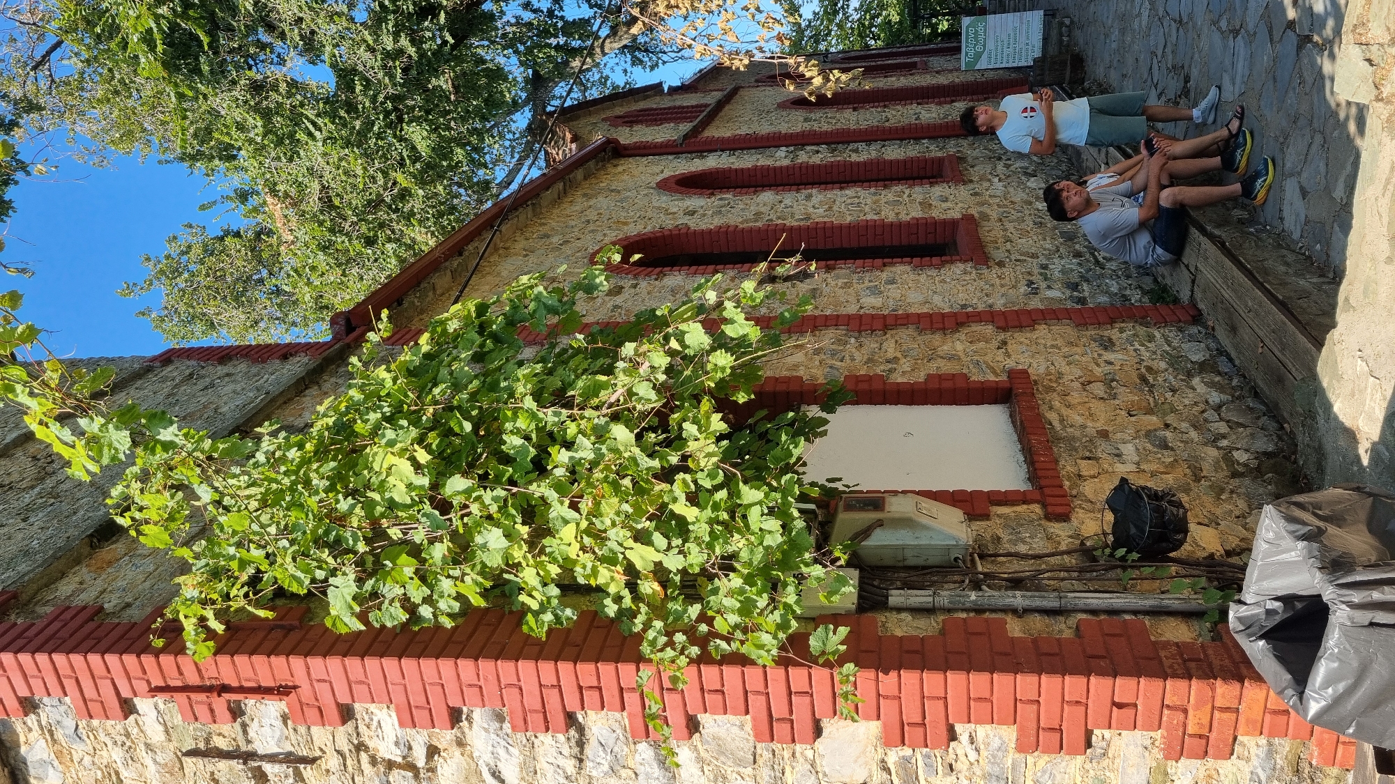 The image depicts the exterior of a stone building with red brick accents, likely a historical structure or church, located in the village of Palaios Panteleimonas, Greece. A lush green vine climbs up the left corner of the building, partially obscuring a white-boarded window and an electrical box. Two people are seated on a wooden bench in front of the building, one man and one woman, both appearing to be looking to their right. A young man stands further to the right, also looking to his right. Behind him, a sign with Greek text is visible, likely indicating a local establishment. The ground is paved with stone, and the scene is bathed in sunlight, suggesting a clear, daytime sky. The overall impression is one of a tranquil, rustic setting.