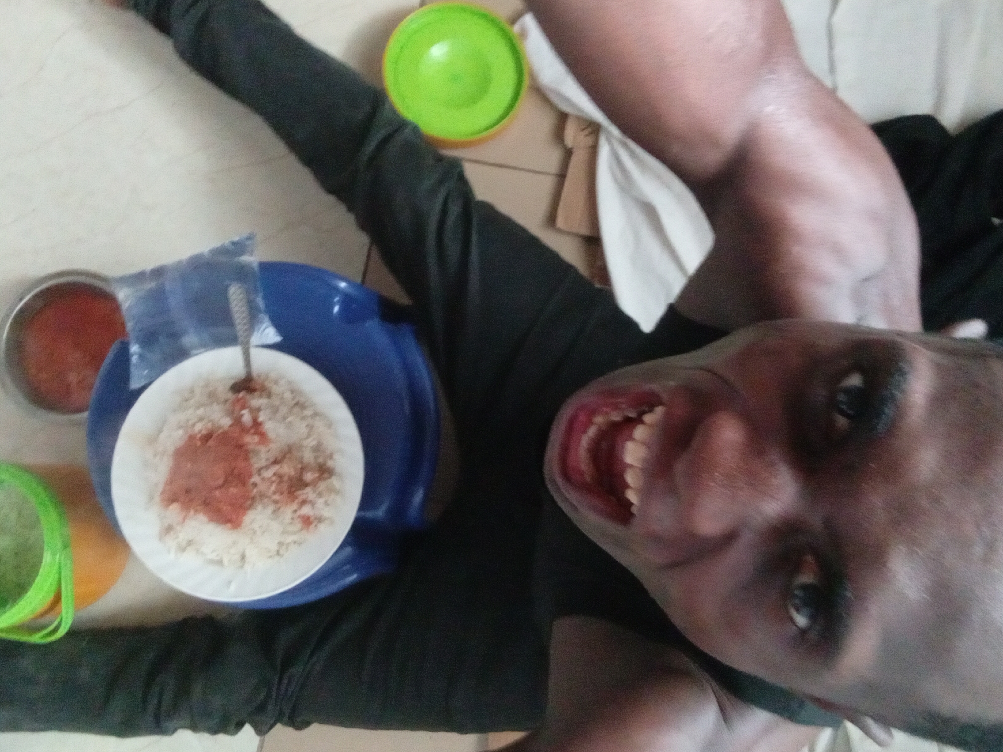 A high-angle view captures a person in a black long-sleeved shirt lying on what appears to be a beige tiled floor. The person's head is positioned in the lower right, tilted towards the camera with their mouth open, revealing their teeth. To the person's left, there is a plate of rice with a red sauce on a blue plate. Additional containers, one green and one red, are placed nearby. The location appears to be indoors. No specific city or location is identifiable.