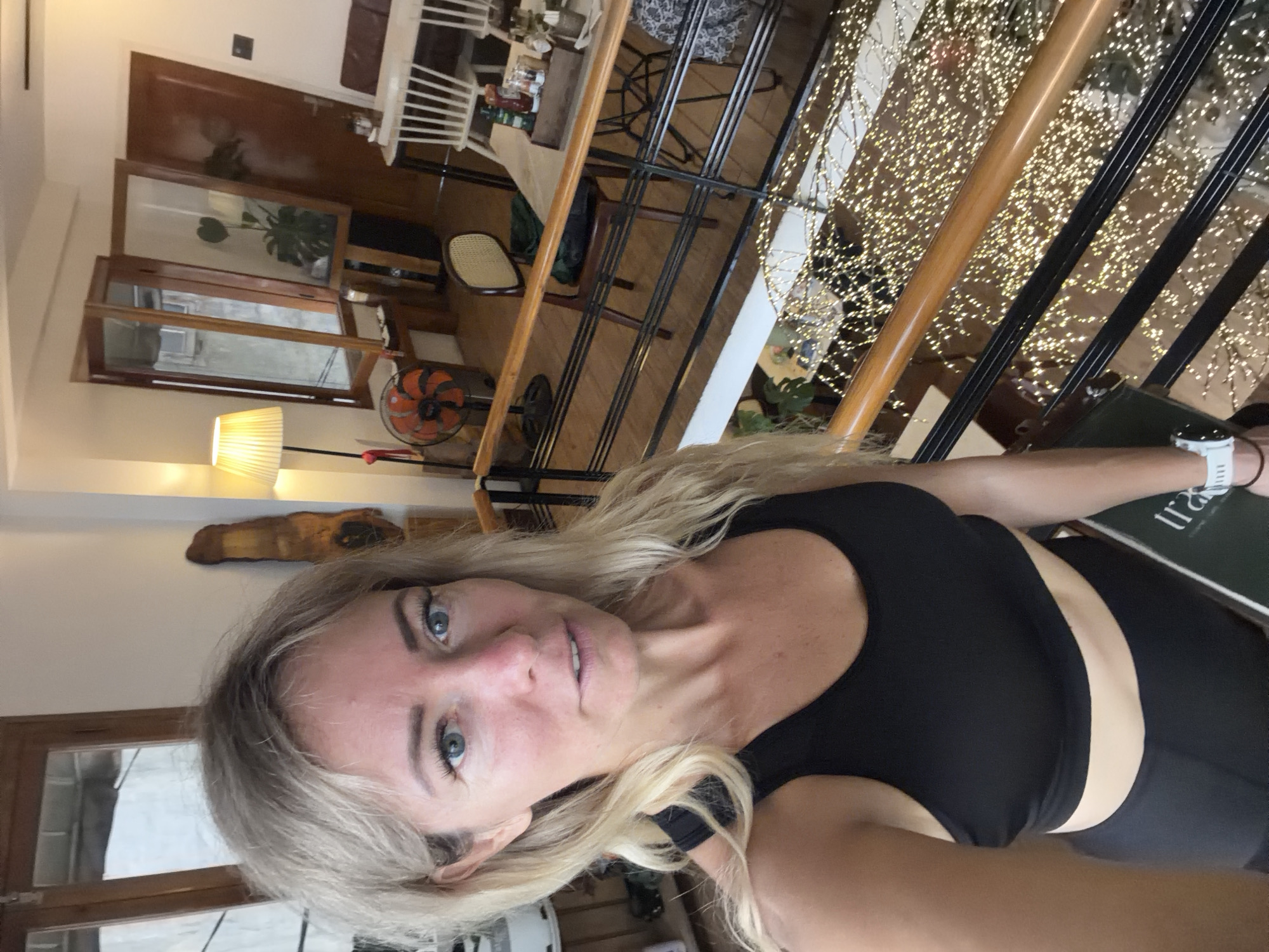 The image shows a blonde woman in a black sports bra and black leggings taking a selfie in an indoor multi-level space, likely a cafe or restaurant, in Da Lat, Vietnam. She is standing on a staircase with a wooden railing. Below her, the staircase is decorated with numerous small, warm-toned string lights. In the background, there is furniture, including chairs and a table with condiments, as well as windows and doors. A fan is visible near a window. The overall ambiance suggests a modern, possibly bohemian, interior design.