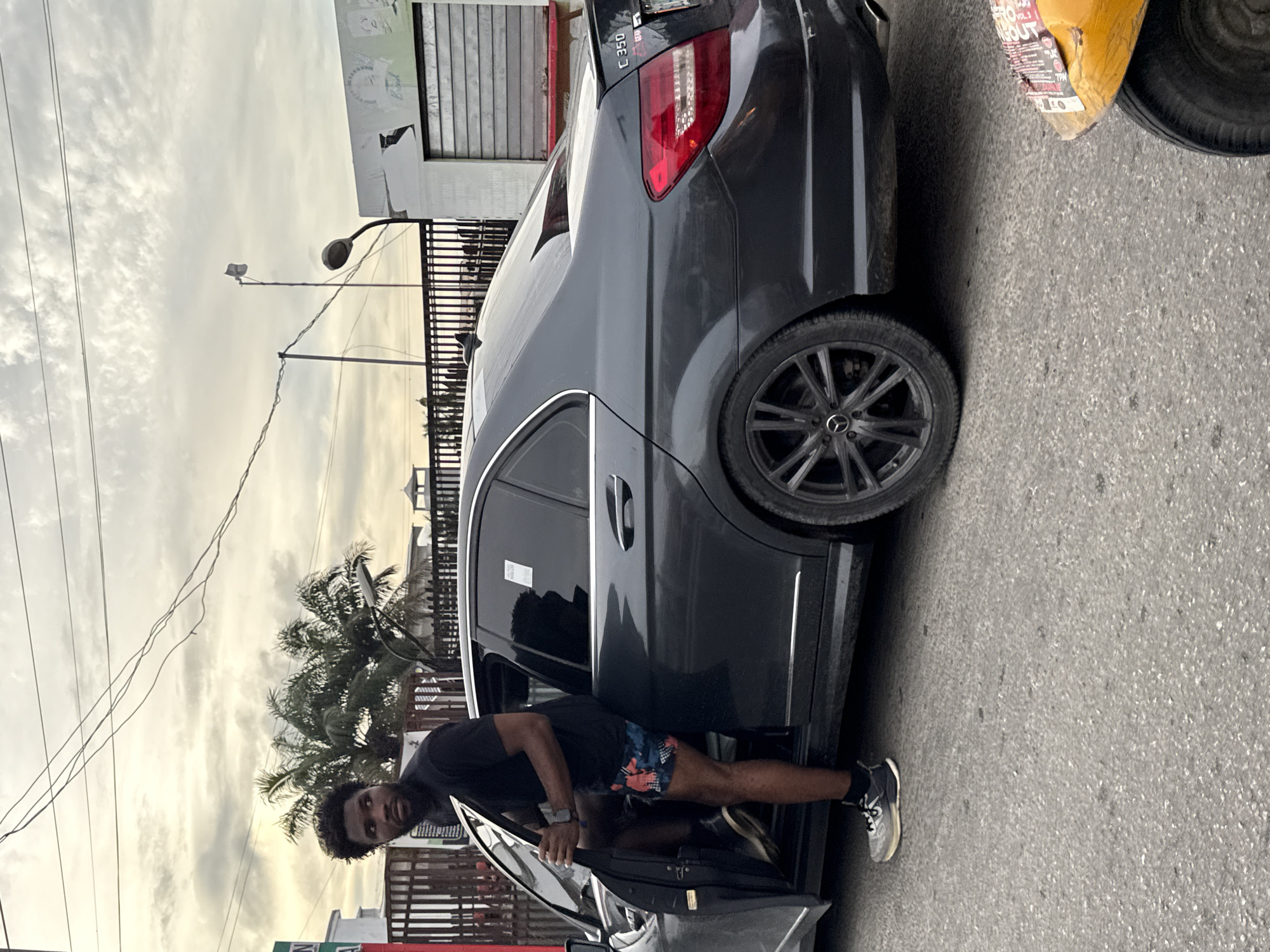 A man is getting into a dark grey Mercedes-Benz C350 parked on a street in Ado, Nigeria. The car is parked next to a commercial building, and a tricycle is partially visible in the top right corner. The man is wearing a dark t-shirt, patterned shorts, and sneakers. Palm trees and power lines are visible in the background. The sky is overcast.