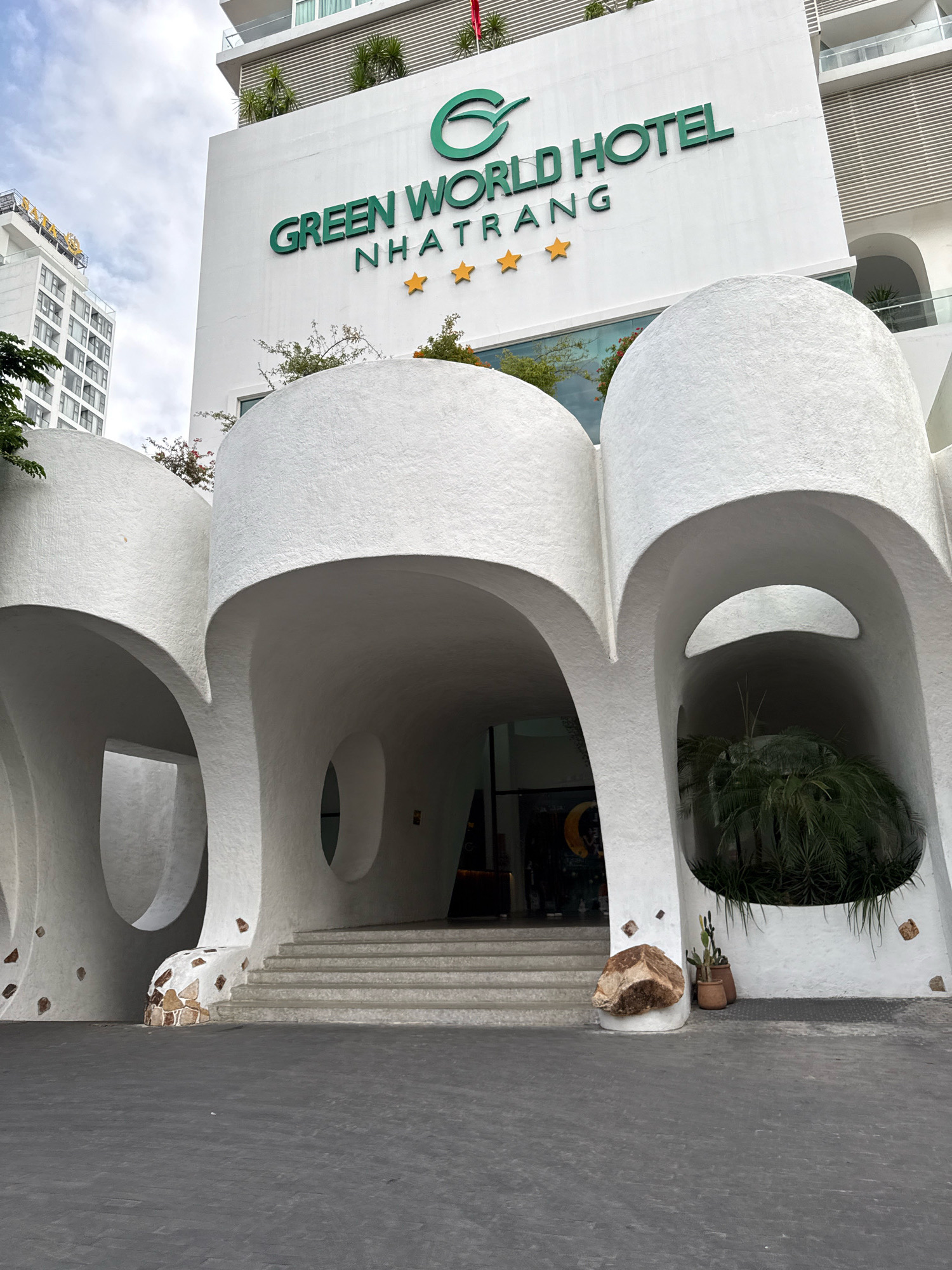 The image shows the exterior of the Green World Hotel in Nha Trang, Vietnam. The hotel has a modern, white, organic architectural design with curved structures and rounded arches. The hotel's name, "GREEN WORLD HOTEL NHATRANG," is displayed prominently in green letters on the white facade, along with three gold stars indicating its rating. The entrance features a set of stairs leading into a covered area. Several plants, including palm trees and cacti, are visible around the entrance and within the architectural elements. In the background, another taller white building, possibly another hotel or apartment complex, can be seen. The ground in front of the hotel is paved with dark gray asphalt. The sky is overcast.