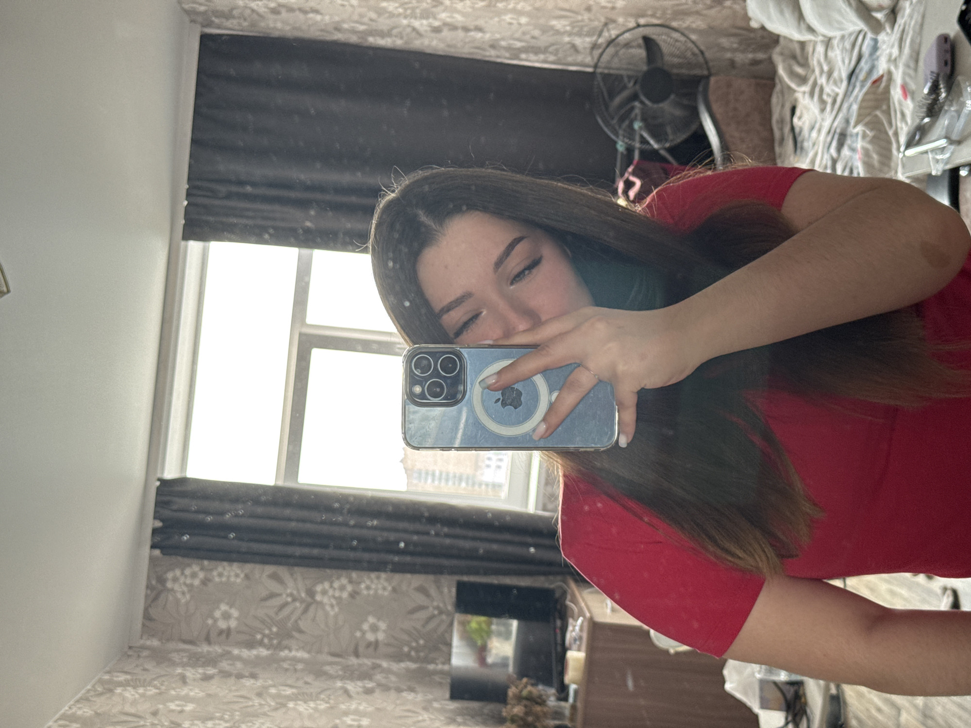 A young woman with long, dark brown hair is taking a selfie in a mirror. She is wearing a red t-shirt. She is holding a blue iPhone in her right hand, covering her mouth and nose. Her left hand is resting on her chest. The background appears to be a bedroom with floral wallpaper. There is a window with dark curtains on the right side of the frame. A television is visible on a wooden dresser to the left. A fan is present behind the woman. The scene is consistent with a residential interior, possibly in a city like Perm, Russia, given the context provided. The lighting suggests it is daytime, with sunlight coming through the window.