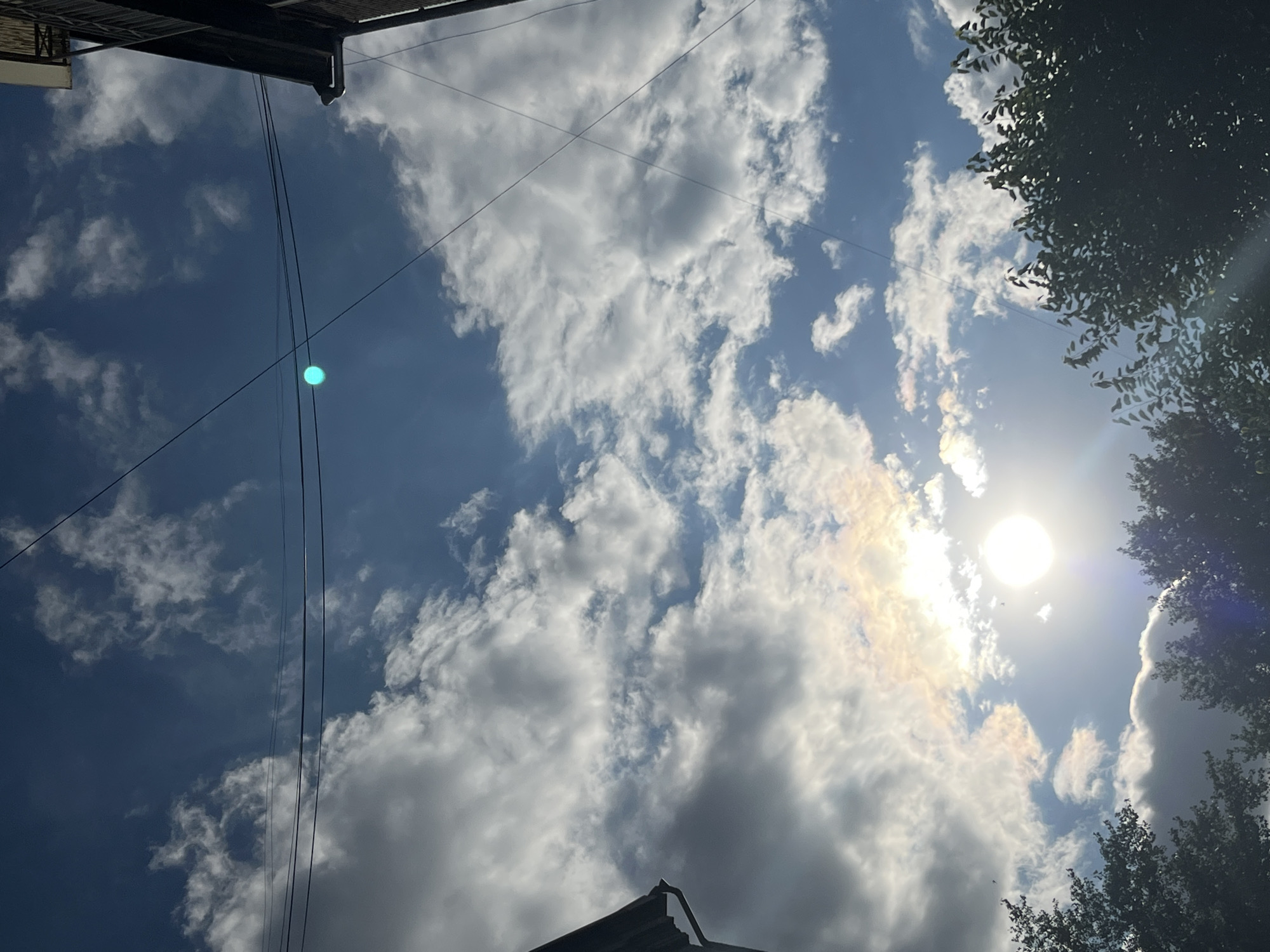 This media file captures a bright, cloudy sky with the sun shining through. The perspective is looking upwards, showing a mix of blue sky and white, fluffy clouds. The sun is visible as a bright, radiant orb, with some iridescent light scattering around it, possibly indicating a halo effect. Several power lines crisscross the sky, suggesting an urban or semi-urban setting. The top of a dark-colored roof or overhang is visible in the upper left corner of the frame, and the dark green foliage of trees frames the right side of the image. The lighting and cloud formations suggest it is daytime, likely late morning or early afternoon. There is a noticeable cyan-colored lens flare in the upper left quadrant of the image. The overall impression is one of a dynamic sky with good weather. Given the provided location context of Dnipro, Ukraine, this scene could represent a typical summer day in the region. No people are visible in the frame, and there is no visible text.