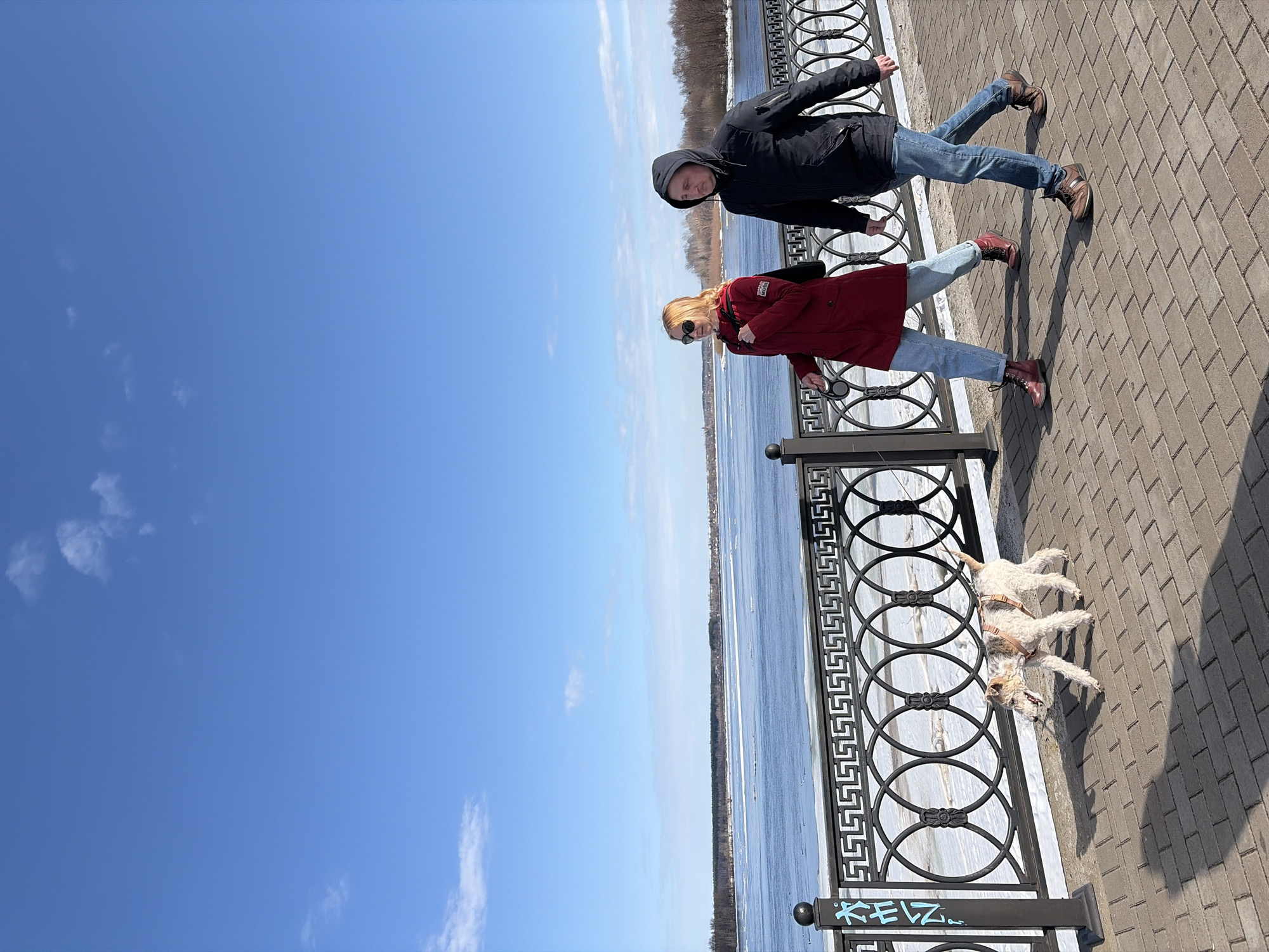 Two people, one male and one female, stand on a paved embankment in Sarapul, Russia, next to a decorative metal fence with geometric patterns. Below, a partially frozen river stretches into the distance. The sky is clear and blue. A dog stands near the fence. The man wears a dark hooded jacket and jeans, while the woman wears a red jacket, sunglasses, and jeans.