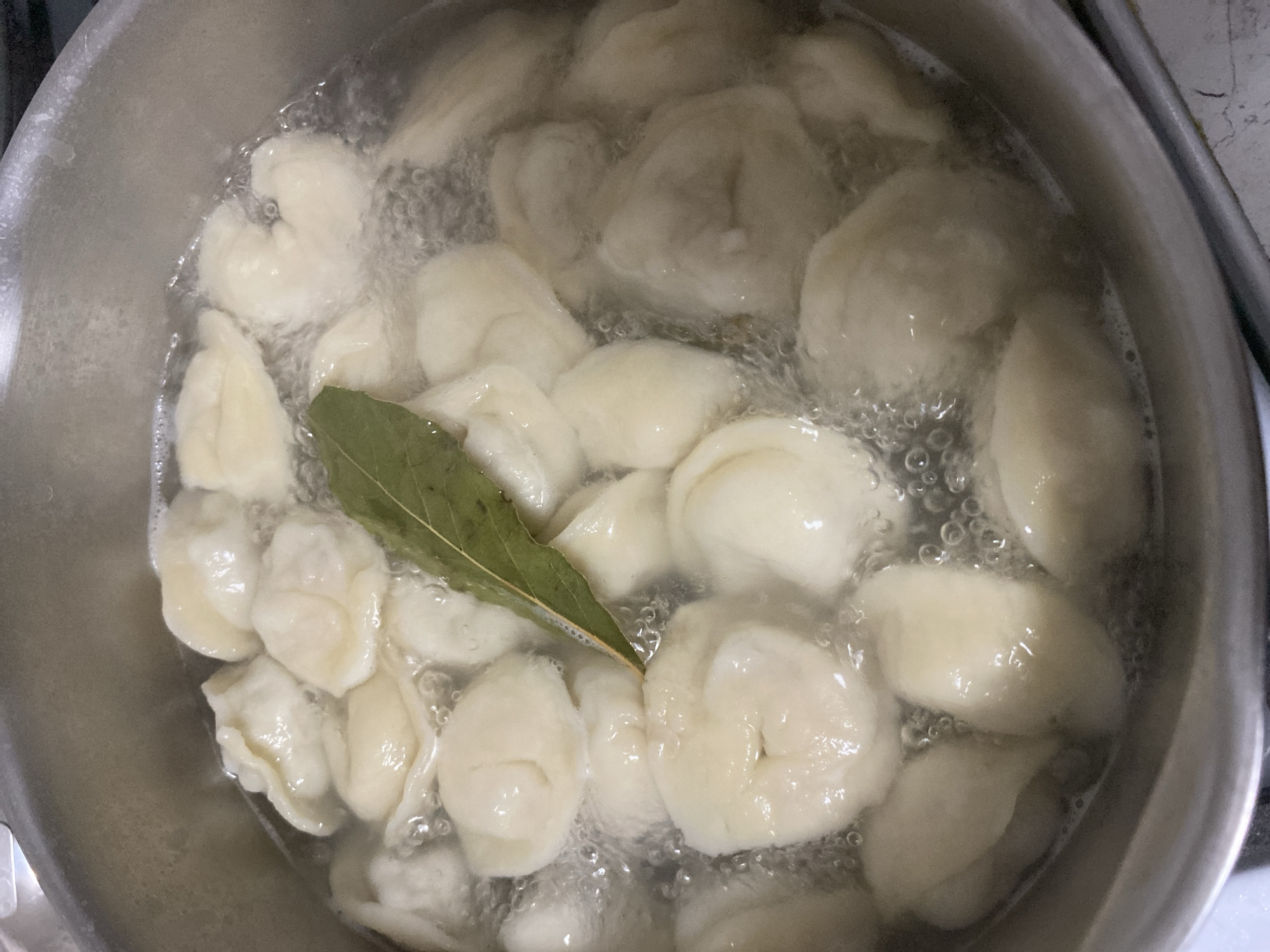 A top-down shot shows a silver pot filled with dumplings boiling in water. A single bay leaf is also in the pot. The dumplings are various sizes and shapes. Bubbles surround the dumplings, indicating the water is actively boiling. The pot sits on a stovetop, with the edge of the stovetop visible. The image was taken indoors. I have no way of knowing the location is Noviy, Russia.