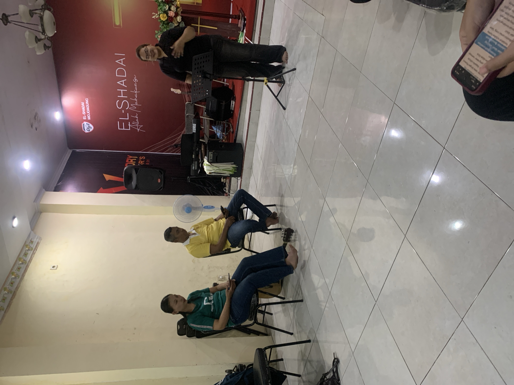 The image captures an indoor scene, likely a church or community hall, in Dukuh Mojo, Indonesia. In the foreground, a person is holding a smartphone, with their hand and part of their leg visible. The mid-ground features a woman in a green shirt and jeans sitting on a chair, looking at her phone, with a guitar leaning against her chair. Next to her, a man in a yellow shirt and jeans is also seated, looking down at his phone. A large blue fan is visible behind them. In the background, a woman in a black shirt and dark pants stands near a music stand, speaking or singing, with a banner behind her that reads "EL-SHADAI MOJOAGUNG" and "EL-SHADAI Allah Mahakuasa." Musical instruments, including a keyboard and speakers, are set up on a stage. The room has tiled flooring and a decorated ceiling with lights. The overall atmosphere suggests a religious gathering or service.