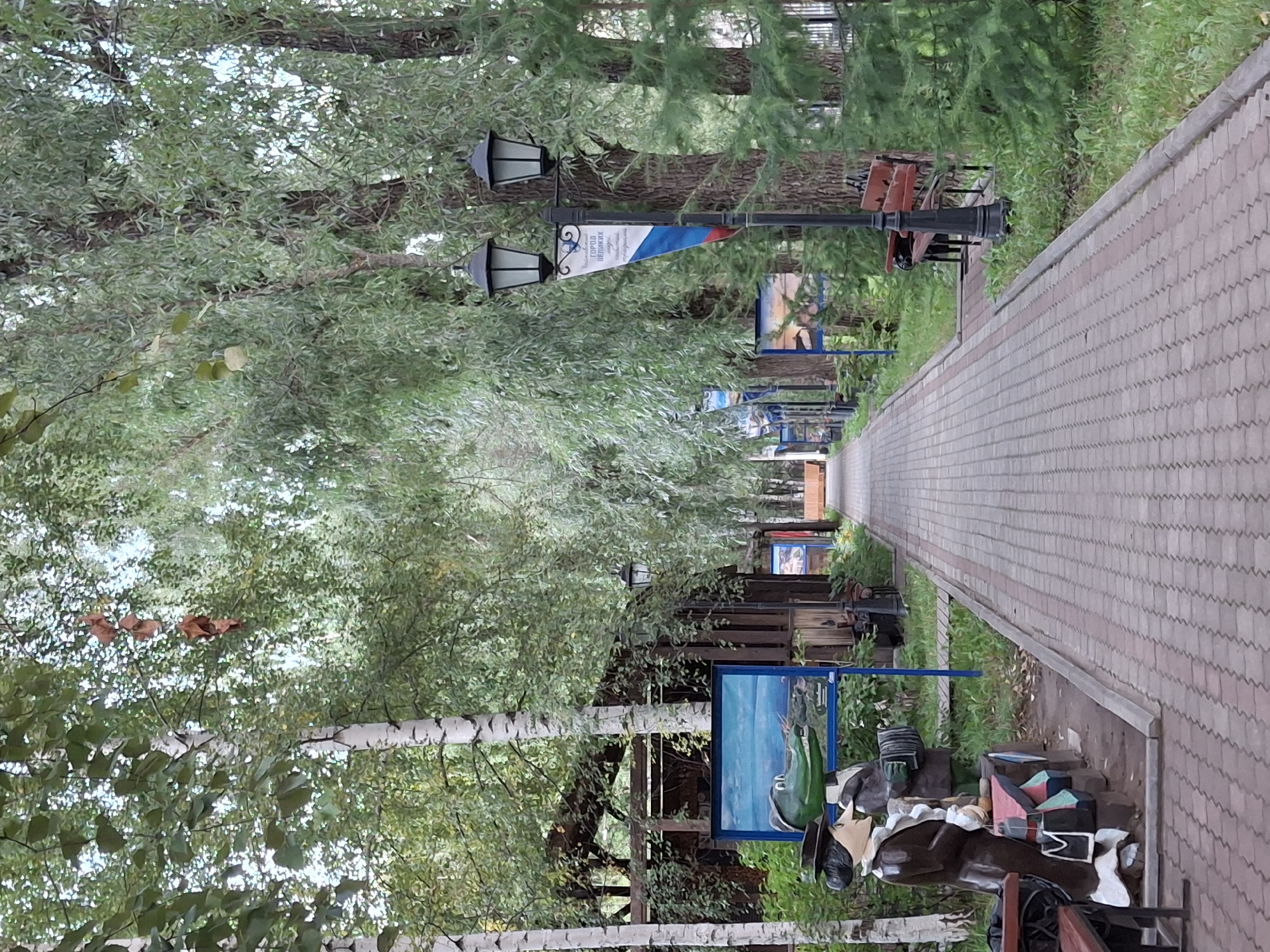 The image depicts a picturesque walkway in Chaikovsky, Russia, likely within a park or recreational area, given the lush greenery and art installations. The pathway is paved with reddish-brown interlocking bricks, bordered by grass and mature trees on either side. The trees, predominantly birch and willow, form a canopy overhead, creating a dappled light effect on the walkway.
On the left side of the path, there are several prominent features. Two wooden sculptures are placed near the foreground: a man in a brown coat and hat, and a green alligator-like creature dressed in a suit and playing an accordion. These figures are positioned next to a wooden structure that appears to be a small pavilion or kiosk, with a painting of a seascape displayed on its side.
Along the walkway, streetlights with black ornate posts and dual lanterns are spaced at intervals. One streetlight in the mid-ground features a triangular banner with Russian text and the national flag colors. Benches are visible on the right side of the path, providing seating for visitors.
Further down the path, the perspective draws the viewer towards a receding vanishing point, with more trees and buildings suggested in the distance. The overall atmosphere is serene and invites a leisurely stroll. The presence of the sculptures and paintings suggests this location is a place of cultural interest or a public art space within the city.