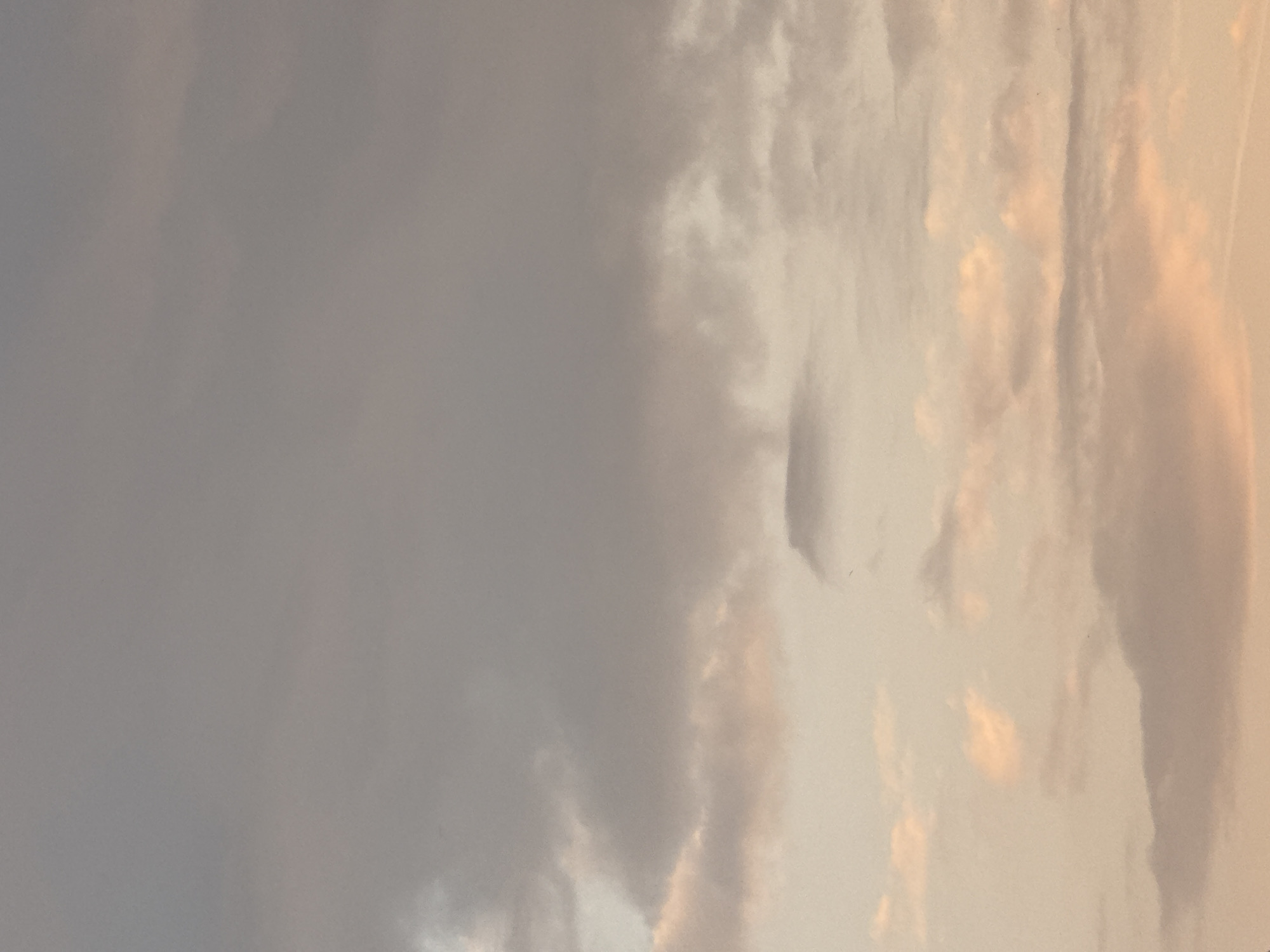 The image shows a cloudy sky. The left side of the image is dominated by gray, overcast clouds, while the right side features clouds tinged with a warm, peach-colored light, possibly indicating the presence of the sun. No people, objects, actions, or interactions are visible. There is no evidence to suggest that this image was taken in Milan, Italy.