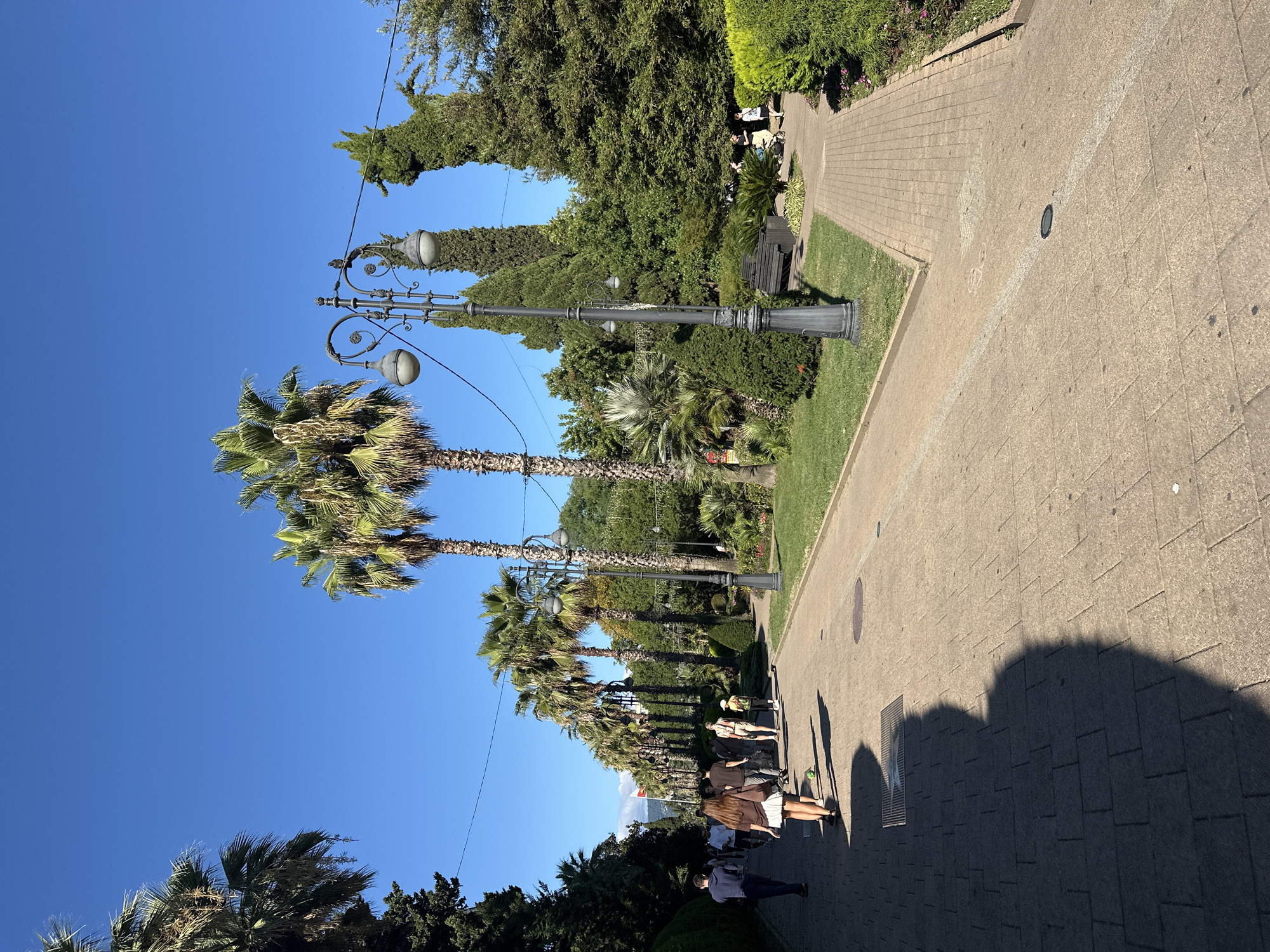 The image depicts a sunny day in a park located in Sochi, Russia, likely on the Black Sea coast, given the presence of palm trees and lush greenery. A wide, paved pathway, made of light brown interlocking stones, runs diagonally from the lower left to the upper right of the frame. People are walking along this path.
In the foreground, a large shadow from an unseen person stretches across the path on the left side. Several individuals are walking away from the viewer, moving towards the right. The group includes a woman in a white skirt and brown top, followed by other people dressed casually. A green soccer ball is visible on the path near the woman.
The pathway is lined with a grassy area to the right, and further on, more palm trees and various other trees and bushes create a dense, green landscape. A decorative black lamppost with two lights stands prominently on the right side of the path, close to a park bench and some smaller palm plants. In the background, more palm trees are visible in a row, creating a sense of an avenue. The sky is clear and bright blue. The overall atmosphere is peaceful and suggests a public space for leisure and recreation.