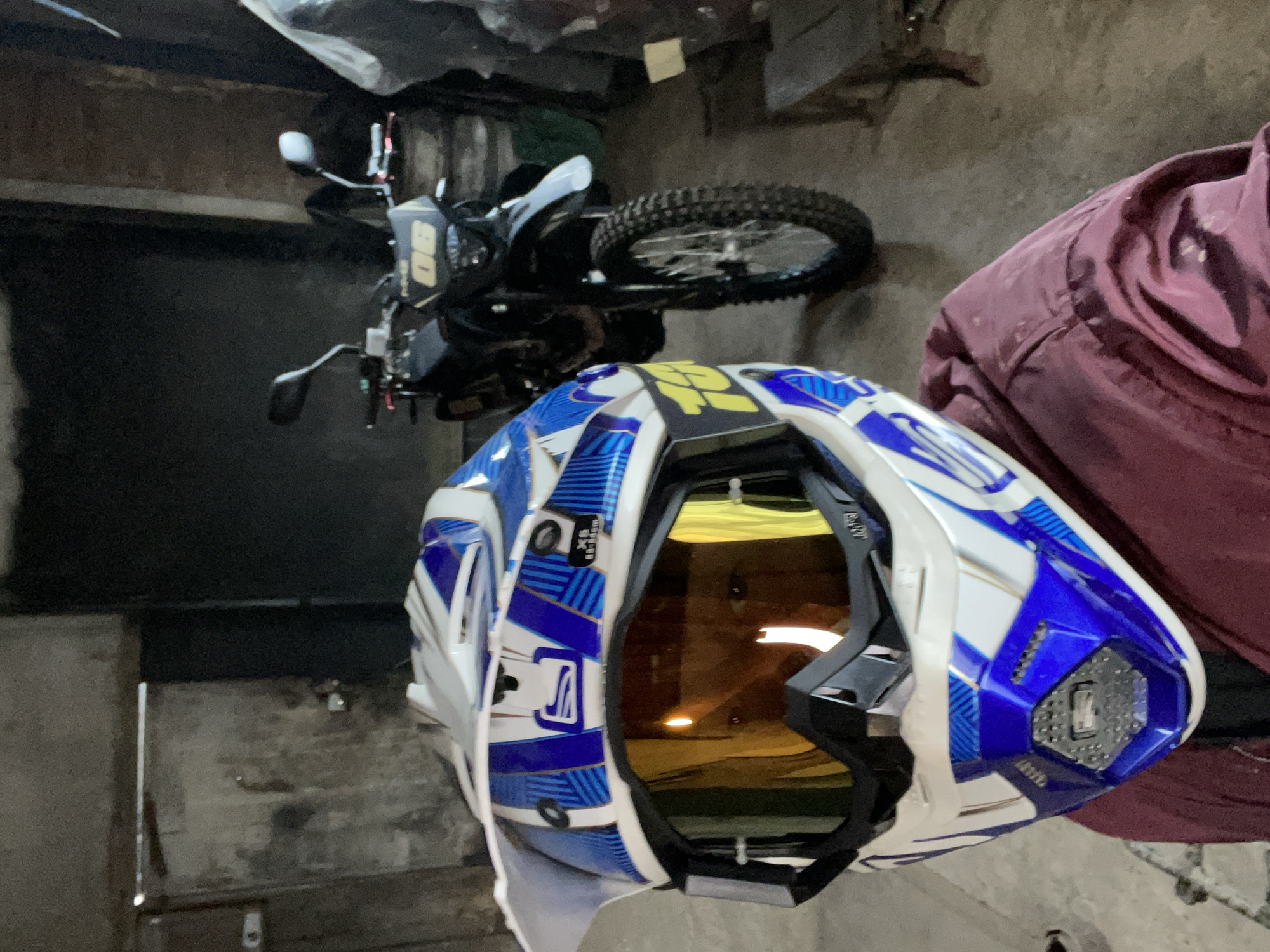 The image primarily features a white and blue motocross helmet with yellow accents, positioned prominently in the foreground. The helmet displays intricate blue geometric patterns over its white shell, with a yellow "100%" logo visible on its top. Integrated goggles, featuring an amber or yellow-tinted visor, are visible through the helmet's opening. A size label "XS 53-54cm" and some additional illegible text (possibly a brand name like "LEATT") are present near the goggle strap.
In the background, positioned vertically due to the image's orientation, is a dark dirt bike or dual-sport motorcycle. Visible parts of the motorcycle include a front wheel with a knobby tire, handlebars, mirrors, and a yellow number plate displaying the number "90" (or possibly "OG"). To the right, a crumpled piece of maroon or burgundy clothing is partially visible.
The scene appears to be an indoor setting, likely a garage or storage area, characterized by rough, dark concrete walls and a similarly textured floor. The lighting is dim, contributing to a utilitarian atmosphere. No specific activity or event is taking place; it is a static shot of equipment. The location context provided is Пастухово, Russia.