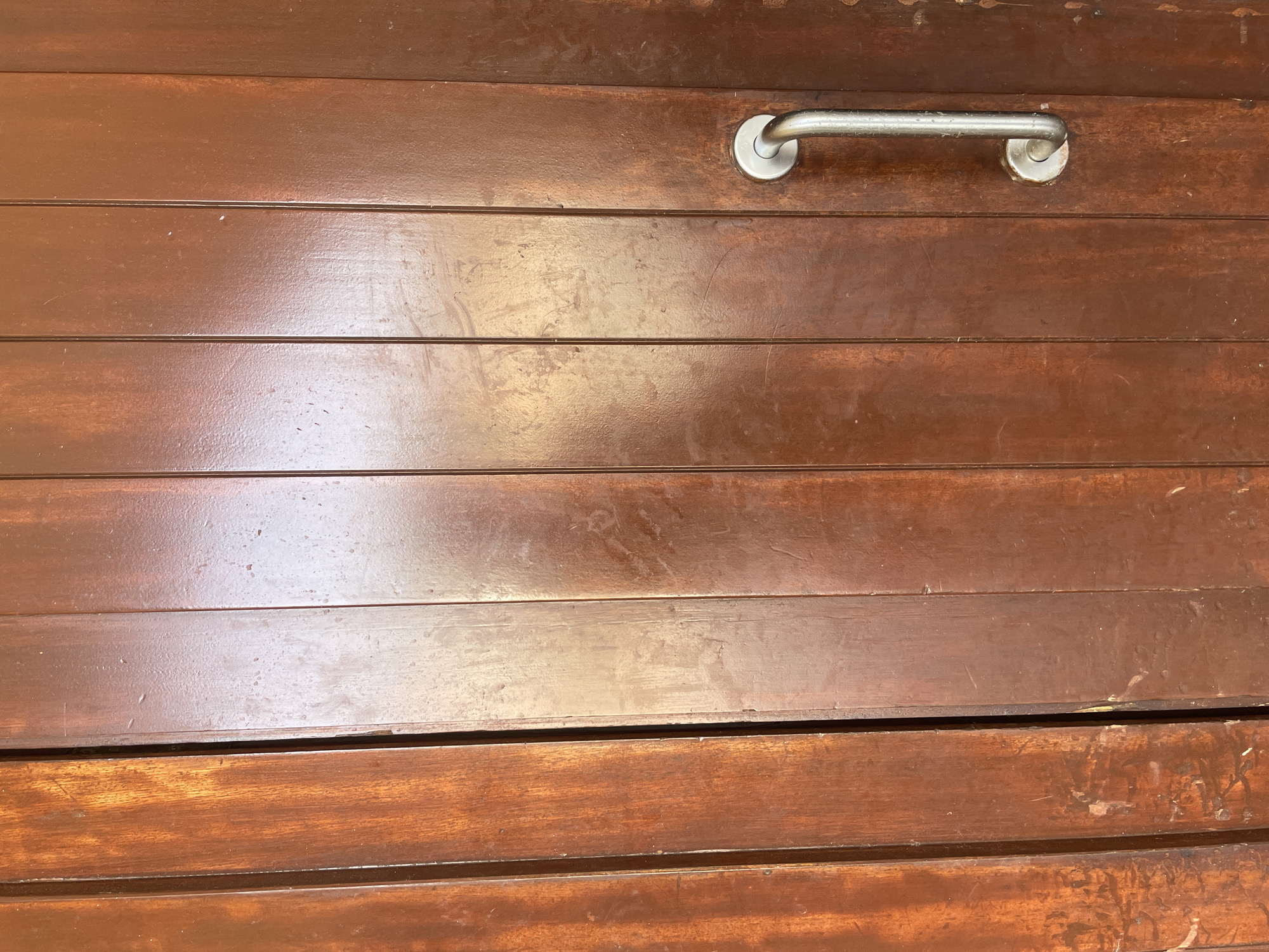 The image shows a close-up of a wooden door with vertical planks. A silver handle is attached to the right-hand side of the door, positioned on a section of planks that is a darker shade of brown than the planks on the left. The wood appears to be varnished and shows some wear, with scuff marks and lighter patches visible on its surface. The lighting creates reflections on the glossy surface of the wood. The scene is devoid of any people or actions. The location of this door in Santanyí, Spain, is not discernible from the visual information provided in the image.