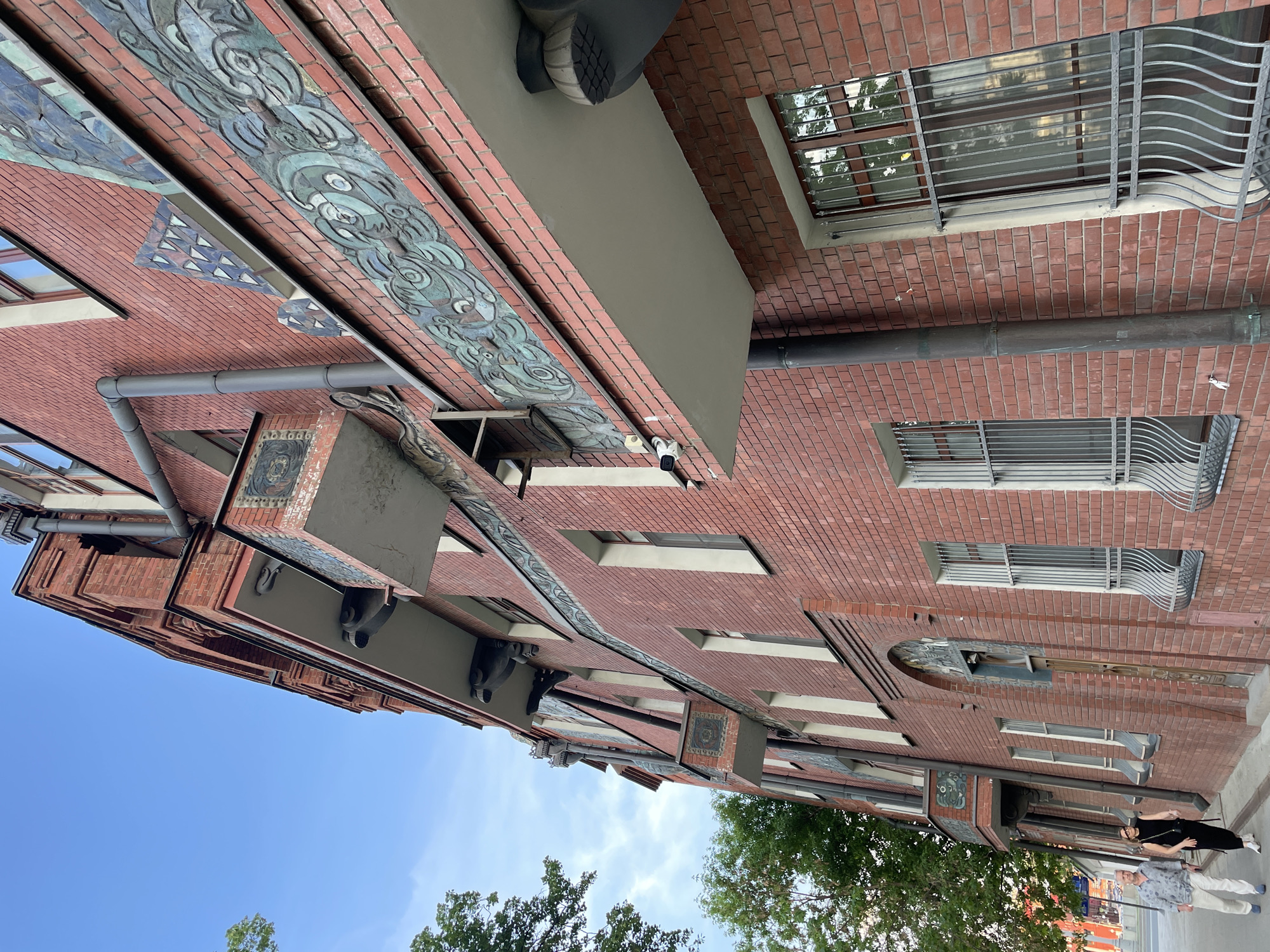 Here's a description of the image:
The image presents the exterior facade of a brick building, likely an architectural landmark. The building is constructed predominantly from red bricks and features decorative elements, including a band of patterned ceramic tiles with blue and green motifs near the top. The windows are rectangular, some with metal security bars. Grey conduits and metal attachments run along the facade. A person is walking on the sidewalk in front of the building. The sky is blue with some clouds.