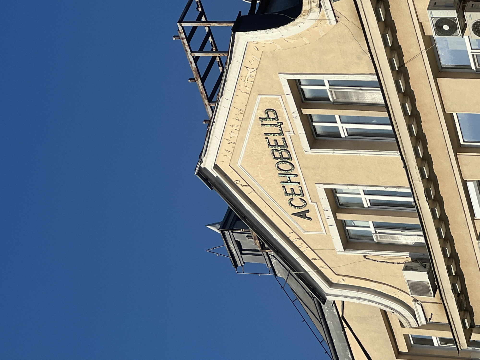 The image shows the facade of a building under a clear blue sky. At the top of the building, a sign with Cyrillic lettering is visible. The letters appear to be in a decorative, possibly illuminated, style, and spell out "АСЕНОВЕЦ". The building itself is painted a light yellow or beige color and features architectural details such as cornices and window frames. There are multiple windows visible, some of which are partially open. Air conditioning units are mounted on the exterior walls. In the background, a dark-colored roof and a section of a metal structure are visible. The overall impression is of an older building in an urban setting.