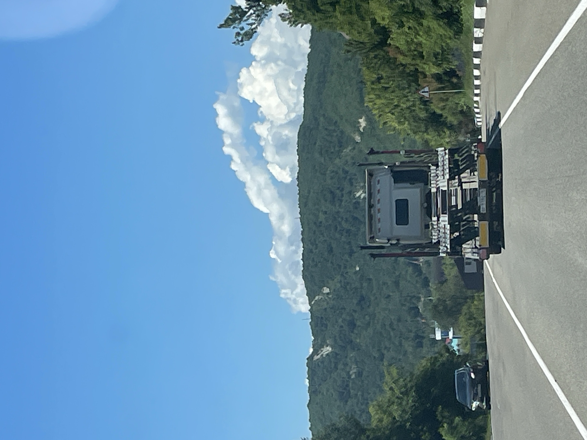 This is a vertically oriented, full-shot, outdoor, daytime photograph taken from a vehicle moving on a highway. The highway is paved with asphalt and has white lane markings. In the foreground, a flatbed truck, seen from the rear, is traveling on the right lane. It is loaded with what appears to be construction equipment or materials. To the left, in the opposing lane, a black sedan is visible. The highway is bordered by a concrete barrier with black and white markings on the right side. Dense green trees line the side of the road. In the background, a large, verdant mountain range dominates the landscape. The sky is bright blue with scattered, fluffy white clouds. The overall scene suggests a journey through a scenic, possibly rural, area. The location is Lermontovo, Russia, on the highway.