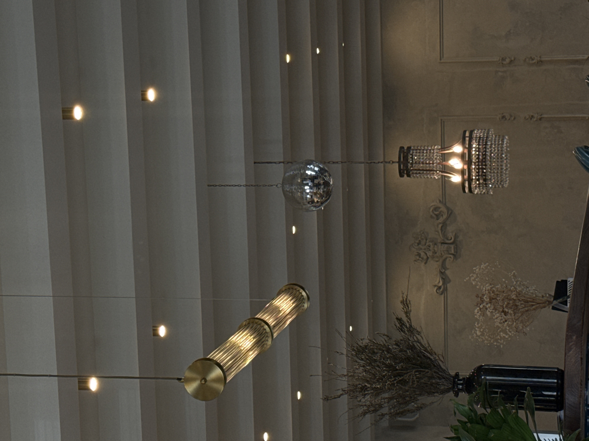 This image captures an eclectic interior space, shot from a low angle looking upwards. The dominant feature on the left is a textured ceiling or tall wall paneling in muted grey tones, featuring embedded, circular spotlights emitting warm light at various intervals.
Three distinct suspended light fixtures are prominent: a classic, faceted disco ball hanging centrally; a cylindrical chandelier composed of golden/brass tubes housing illuminated bulbs, positioned below and to the left of the disco ball; and an ornate, rectangular chandelier adorned with multiple layers of clear, crystal-like elements and internal warm lights, situated to the right of the disco ball. These fixtures are suspended by chains or wires.
To the right, a textured wall with classical decorative molding is visible, suggesting an older architectural style contrasted with modern elements. Below the ornate chandelier, two bunches of dried floral arrangements are displayed. One appears light-colored and airy, while the other is composed of darker, dried grasses or reeds, housed in a dark, cylindrical bottle or vase. A cluster of green foliage is partially visible at the very bottom right corner.
The scene suggests an indoor setting, likely a cafe, restaurant, or event venue, lit by artificial light, implying an evening or nighttime ambiance. No people are visible, and there is no discernible text. The image composition emphasizes the decorative lighting and textural elements of the room.