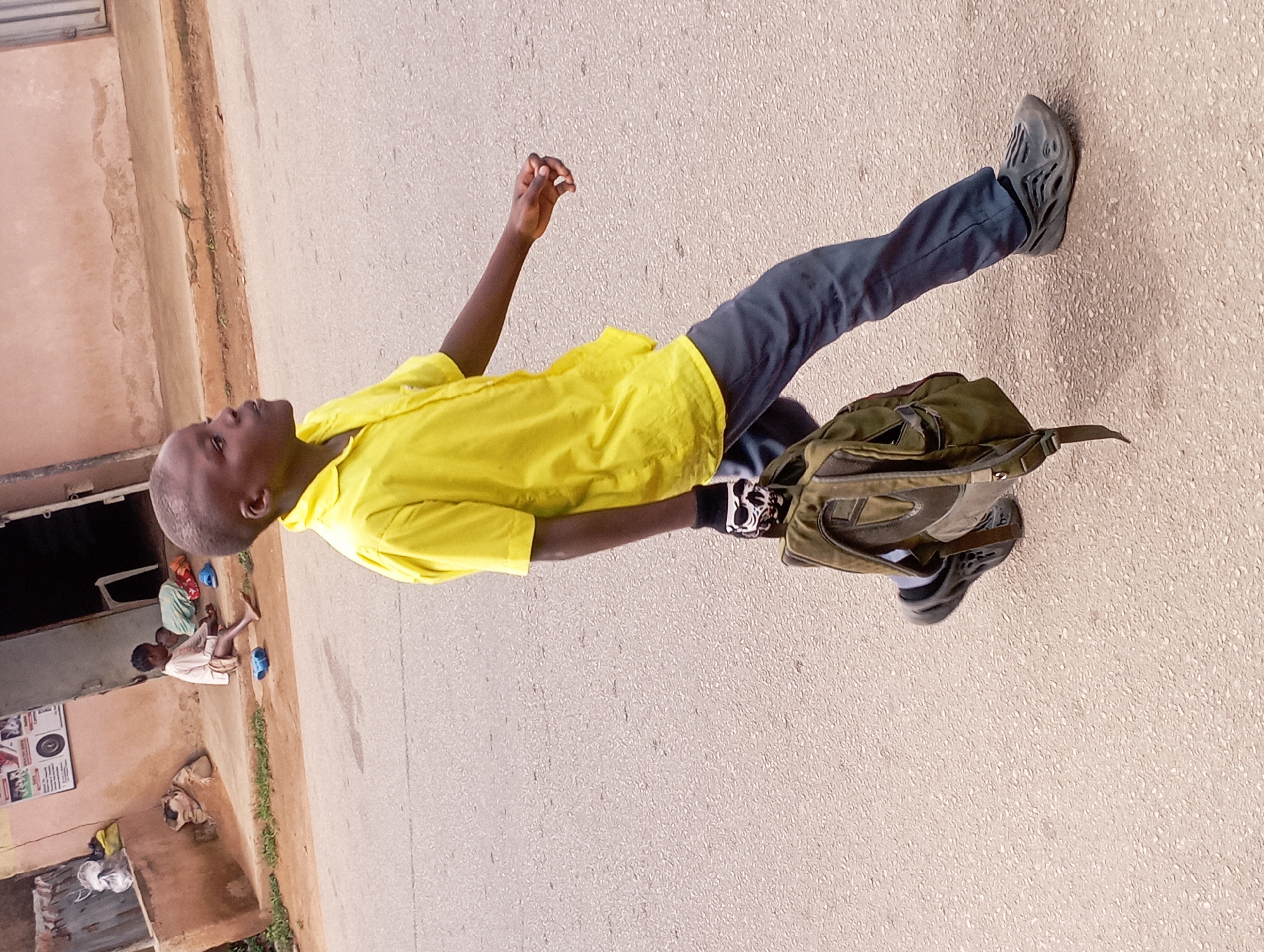 The image depicts a young boy in a yellow shirt and dark pants walking along an asphalt road in Jos, Nigeria. He carries a green backpack in his right hand, wearing a black glove with a skull design. In the background, two younger children are seated on a concrete step in front of a building with a poster on the wall. The environment appears to be a residential or commercial street with buildings and a dusty ground.