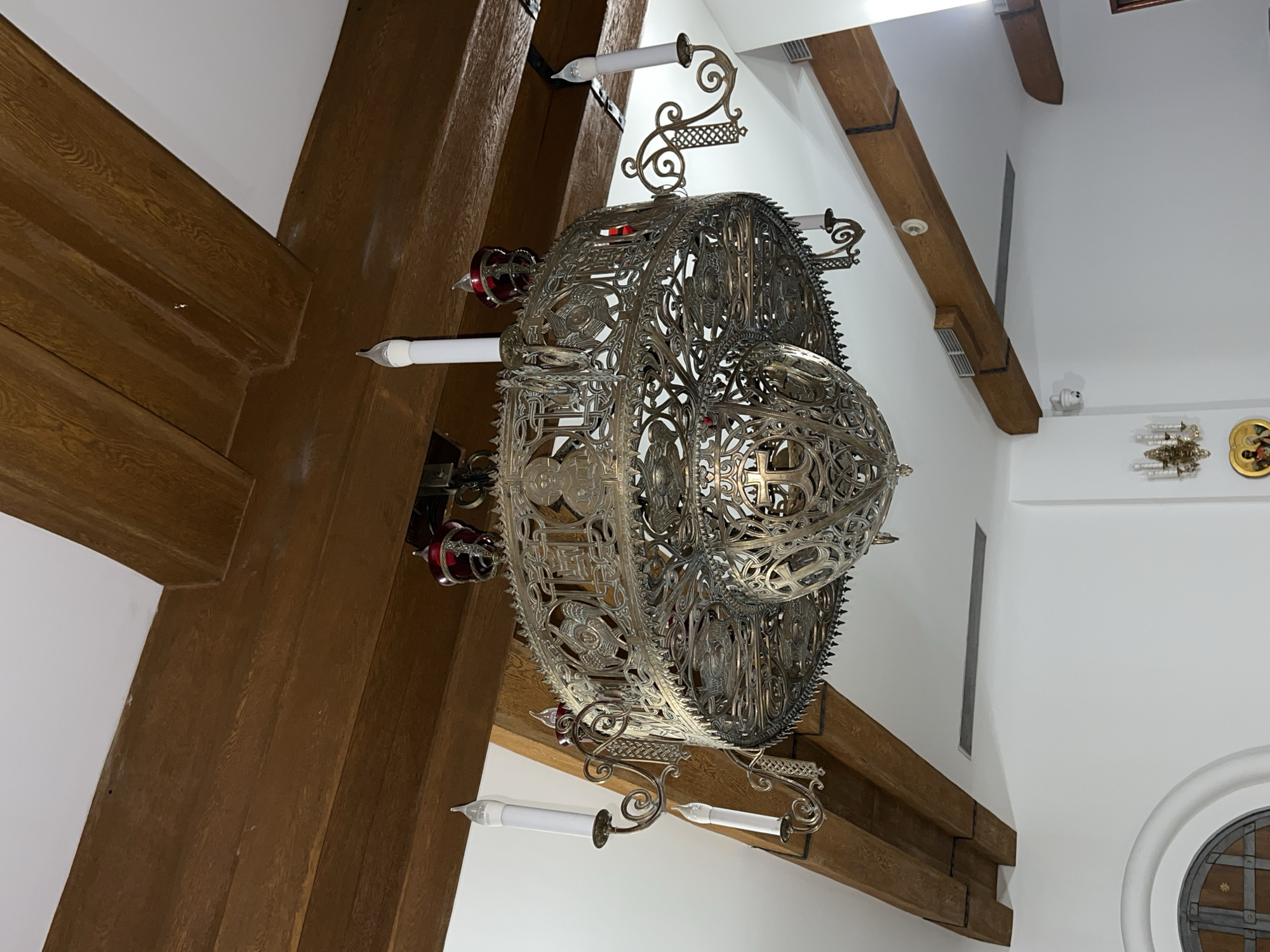 The image depicts an ornate, metal chandelier hanging from wooden beams in an interior space. The chandelier features intricate latticework and candle-like lights. The wooden beams are dark brown and cross each other. The walls are white, and there are other light fixtures and decorative elements visible in the background. There is no indication in the image that this location is in the city of Khimki, Russia.