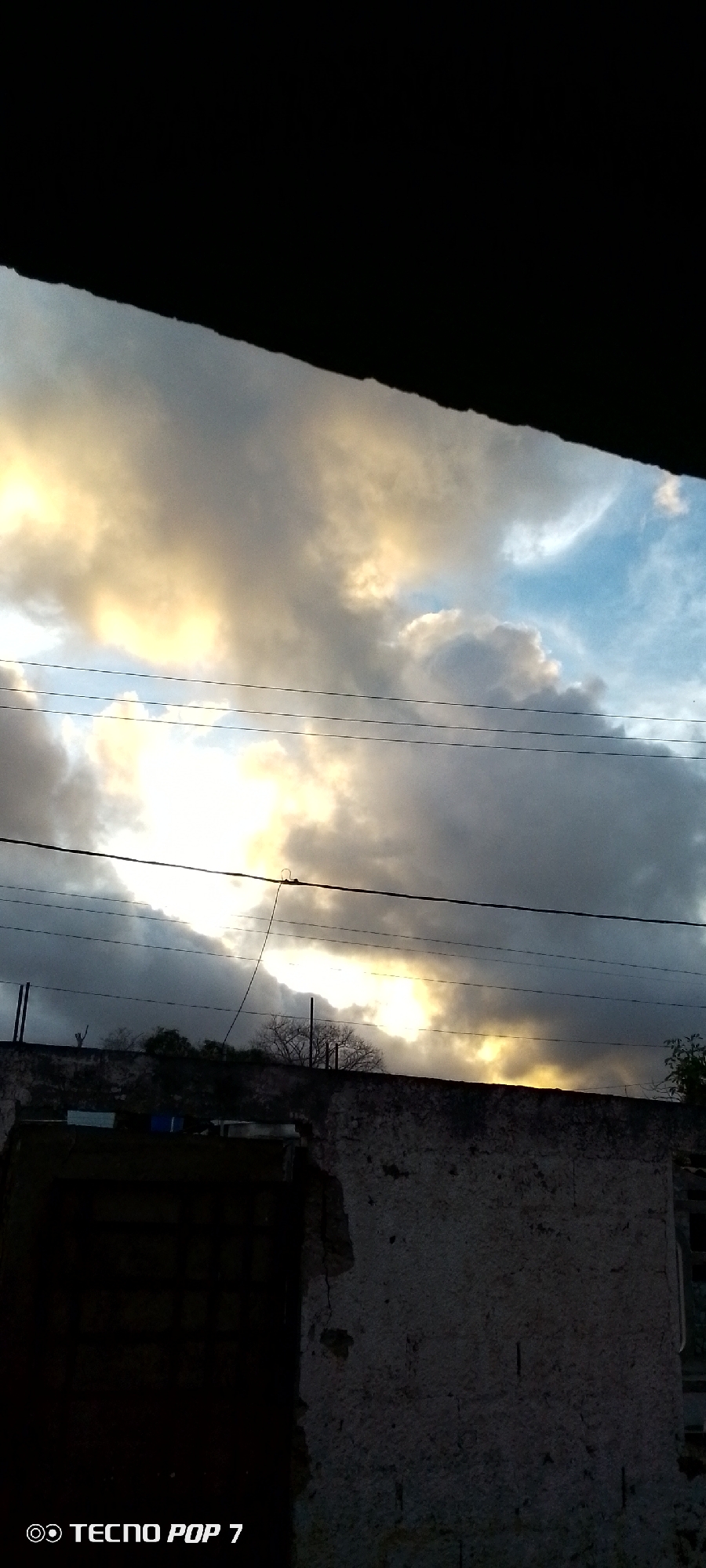 The image captures a dramatic sky, likely during sunrise or sunset, as viewed from a low angle, possibly from beneath an overhang. The top of the frame is dominated by a solid black area, suggesting a roof or structure directly above the viewer.
The sky is filled with thick, textured clouds displaying a striking contrast of colors. Sections of the clouds are brightly illuminated with warm, golden-yellow light, indicating the sun is low on the horizon and breaking through the cloud cover. Other parts of the clouds are a darker grey, creating depth and shadow. Patches of pale blue sky are visible in between the clouds.
Multiple thin, dark utility wires or power lines stretch horizontally across the middle section of the image, intersecting the cloudy sky. One wire in the center appears to have a connection point or loop.
Below the sky and wires, the upper edge of a weathered building or wall occupies the mid-ground. The surface appears to be rough, possibly concrete or stucco, showing signs of wear, cracks, and some damage. Behind this wall, silhouetted against the bright clouds, are the sparse, bare branches of what seem to be deciduous trees or bushes. A dark, indistinct rectangular shape is visible on the left side of the wall, potentially a window or opening.
The overall scene conveys a sense of the transitional light of dawn or dusk. There is no visible human activity. In the bottom left corner, the watermark "O TECNO POP 7" is present.