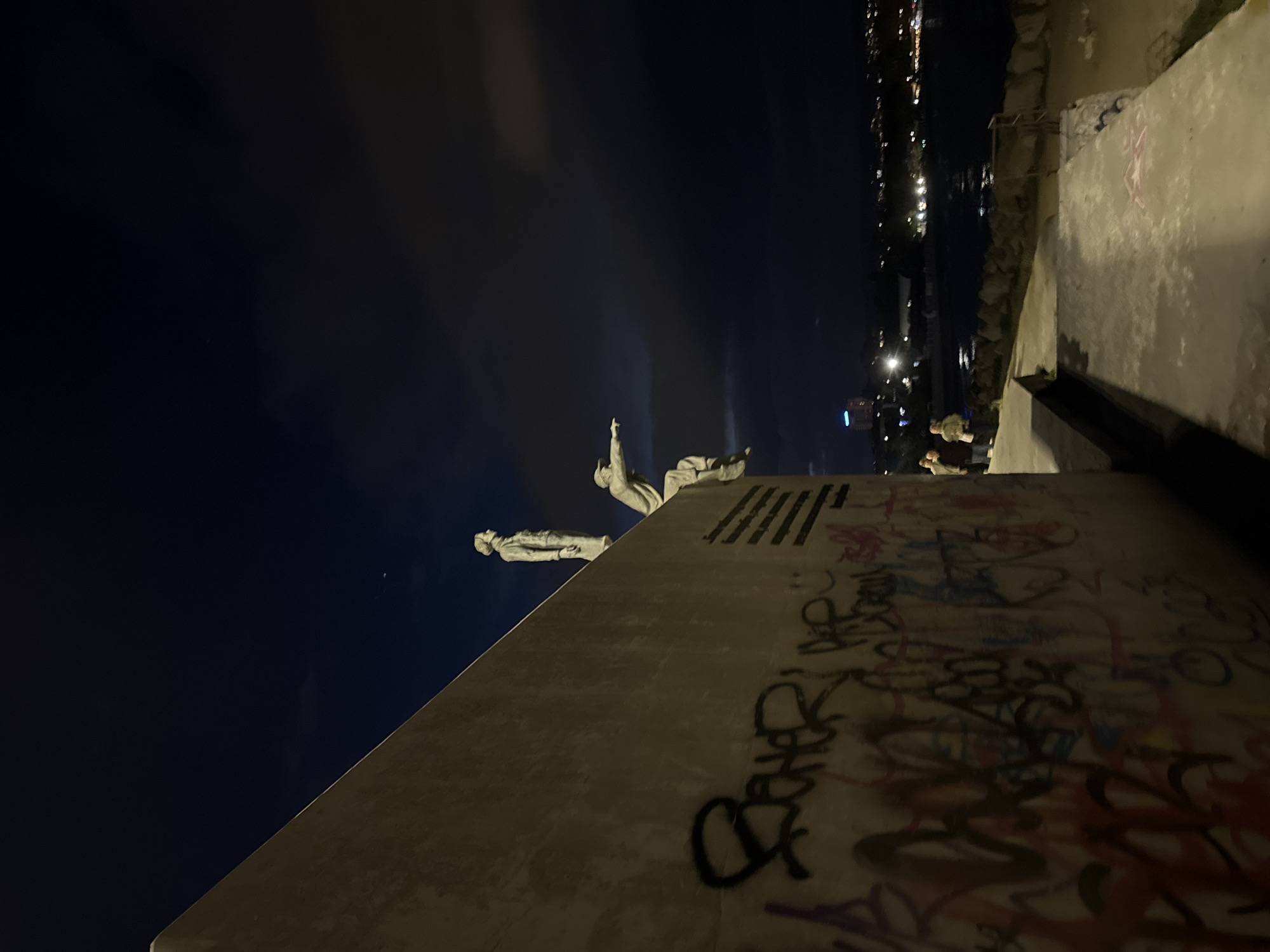 At night, two sculptures stand on a pedestal covered in graffiti in the city of Unknown. Behind the pedestal, a river flows towards a city with illuminated buildings. Near the river, two people are standing.