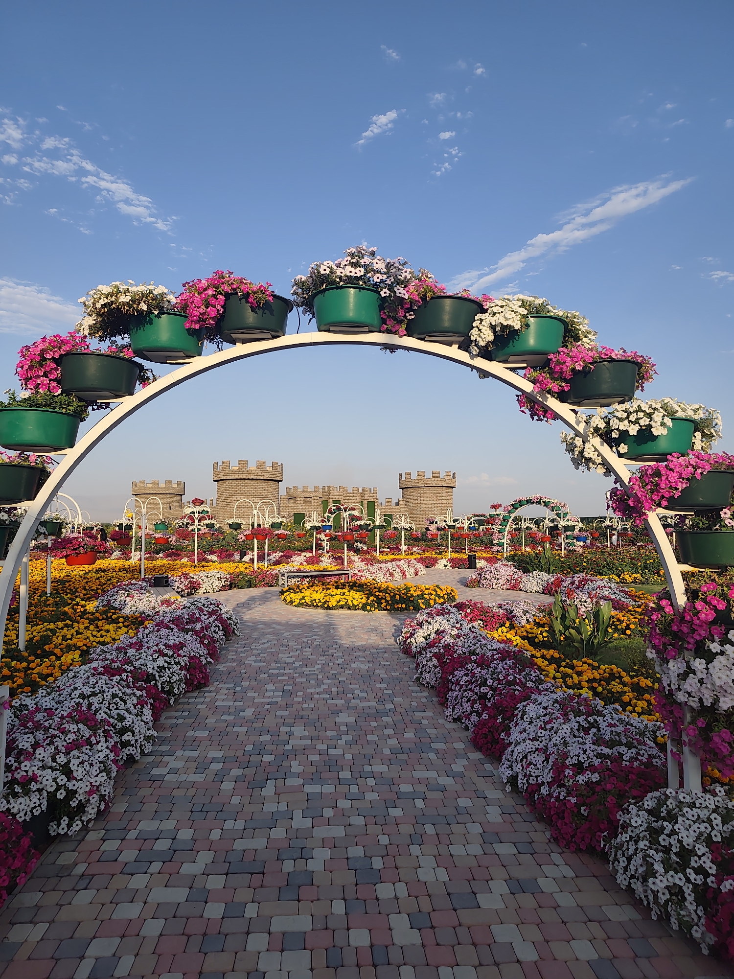 This image depicts a vibrant flower garden with a walkway leading towards a backdrop of castle-like structures. The foreground features a curved archway adorned with numerous green planters overflowing with pink and white flowers. The walkway itself is paved with multi-colored rectangular stones, bordered by extensive flower beds. These beds are densely planted with a profusion of pink, white, and yellow flowers, creating a colorful and lush landscape.
In the mid-ground, several smaller arched structures, also decorated with flowers, punctuate the garden. Behind these, the castle-like structures, resembling medieval towers with crenellated tops, stand prominently. The sky above is a clear blue with scattered white clouds. The overall scene is one of a well-maintained and aesthetically pleasing garden. The text provided states this is the city of Khor Virap, Armenia, though the visual elements do not specifically confirm this location.