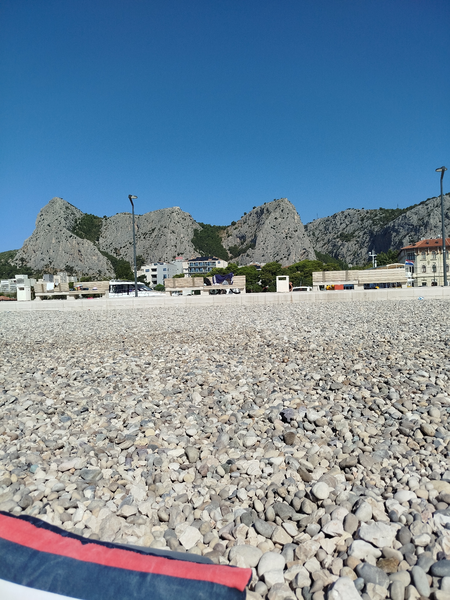 The image captures a sunlit scene of a pebble beach and a promenade in Omiš, Croatia, with the dramatic Biokovo mountains serving as a backdrop. The foreground is dominated by a dense field of grey and beige pebbles. A low, white concrete wall separates the beach from a paved promenade, which features several wooden benches and a trash receptacle. In the middle ground, a modern white boat is docked near the promenade. Beyond the promenade, buildings with white and red facades are visible, interspersed with lush green trees. A tall, dark lamppost stands prominently on the promenade, casting no discernible shadow. The sky is a clear, deep blue. In the lower left corner, a portion of a boat or raft is visible, with a red stripe running along its edge.