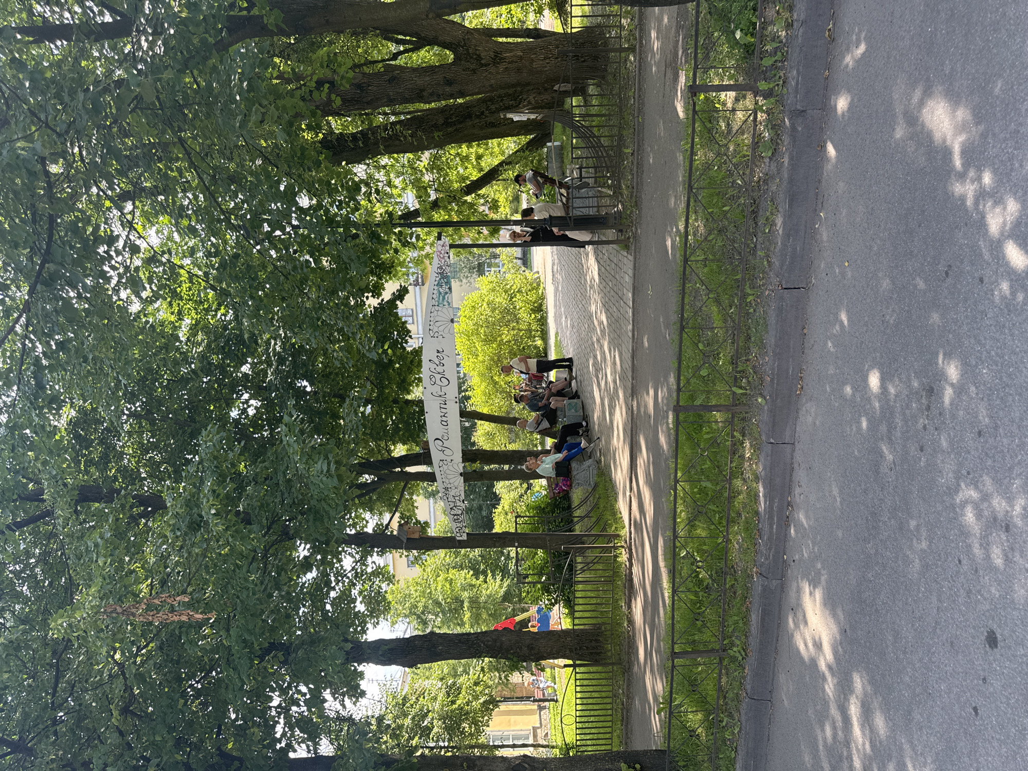 In Metallostroy, Russia, a sunny park scene unfolds. A white banner, adorned with intricate floral patterns and Cyrillic lettering reading "Романтик-Клуб" (Romantic Club), is strung between two large trees, casting a shadow on the checkered pathway below. Several individuals are seated on benches, some engaged in conversation, while others observe their surroundings. A playground structure with red and blue elements is visible in the background, suggesting a family-friendly atmosphere. A dark metal fence delineates the park area from a paved road, where dappled sunlight filters through the overhead foliage. A single birdhouse is mounted on a lamppost.