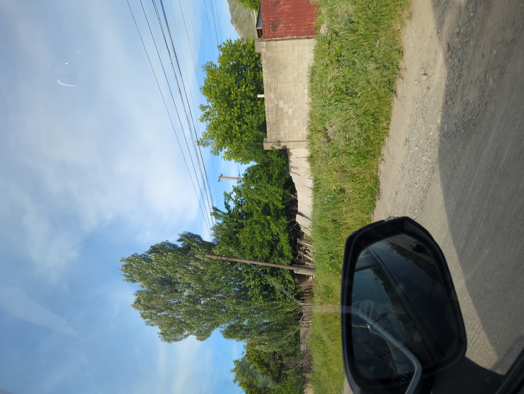 The view from a moving vehicle shows a roadside in Beş-Küŋgöy, Kyrgyzstan. The sky is partly cloudy with power lines running overhead. Green trees line the road, next to a concrete block wall topped with a red roof. A dirt road runs alongside the green vegetation. The car's side mirror is visible in the foreground.