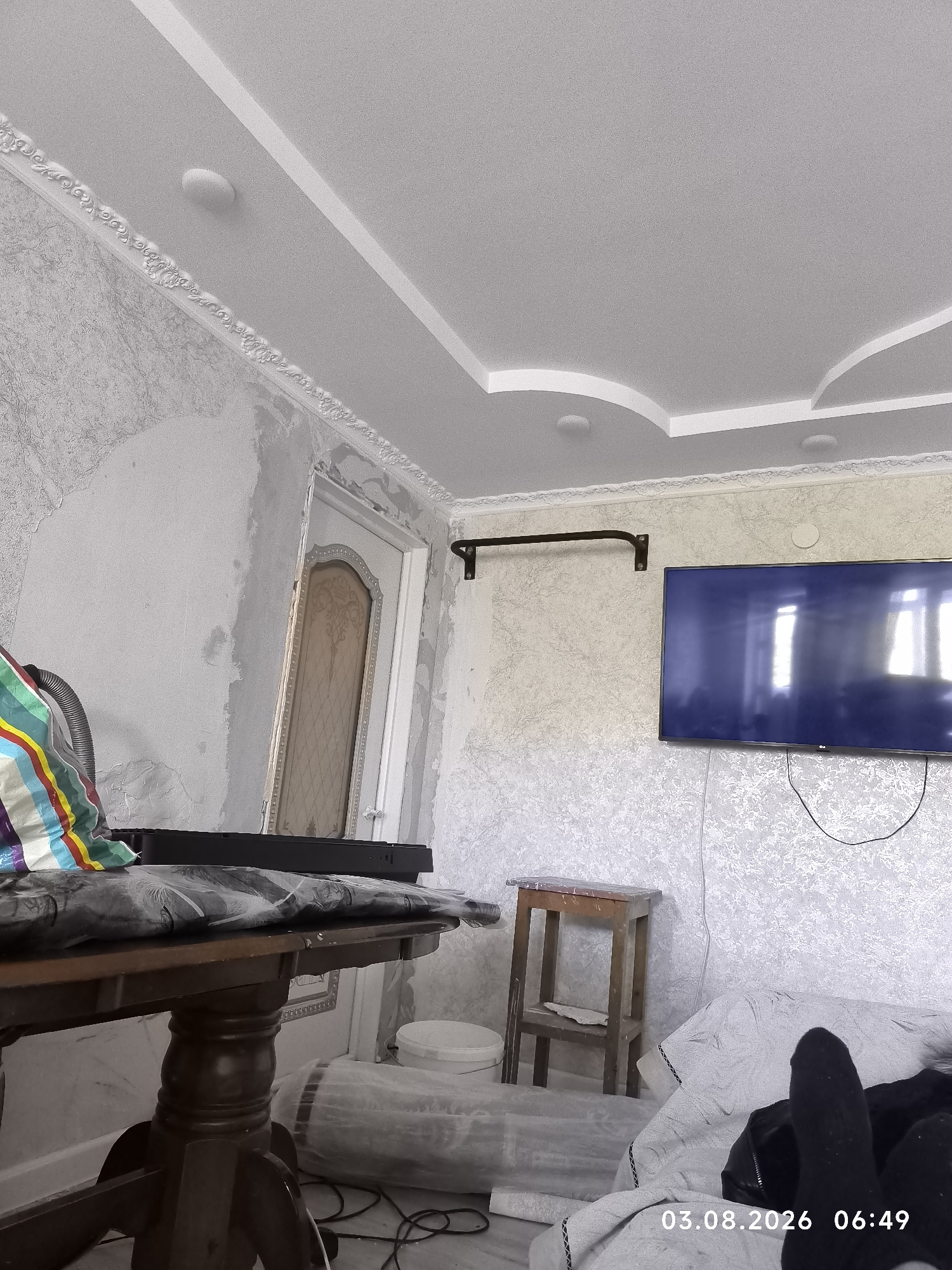 The image depicts an indoor scene, likely a room undergoing renovation or being prepared for one, in the city of Satbayev, Kazakhstan. A dark wooden table with ornate legs is positioned in the foreground, covered with a dark cloth patterned with what appears to be palm leaves. A colorful striped fabric is also visible on the table, along with a vacuum cleaner hose. Behind the table, a white door with decorative panels stands slightly ajar, revealing a glimpse of the room beyond.
The walls are covered in a textured wallpaper, and some areas appear to have been scraped or are awaiting finishing. A large flat-screen television is mounted on the wall to the right, reflecting light from unseen windows. A thin black bar is mounted on the wall above the television, possibly a curtain rod or a pull-up bar. A small wooden stool with a rough-hewn top is placed near the television.
In the lower right corner, a person's feet, clad in black socks, are visible resting on what appears to be a white blanket or bedding. The date and time stamp "03.08.2026 06:49" is superimposed on the image. The overall impression is one of a residential space in a state of transition, with elements of home furnishings and renovation materials coexisting.