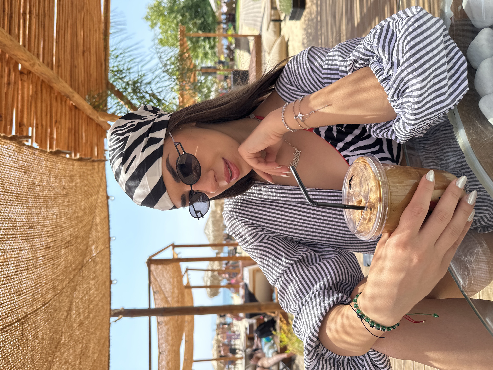 In Ofrynio Beach, Greece, a young woman with long, dark hair and a zebra-print bandana sits at a glass-topped table under a thatched umbrella. She wears a black and white striped open shirt over a zebra-print bikini top, and black oval sunglasses. Her left hand holds a plastic cup with a brown beverage and a black straw. She wears multiple bracelets on her wrists, including one with green beads on her left wrist and a delicate rose-gold chain with charms on her right. A silver necklace with her name, "Anhelika," is visible. The background shows a sandy beach with more thatched umbrellas, lounge chairs, and people enjoying the sun. The overall scene is one of a relaxed beachside vacation.
