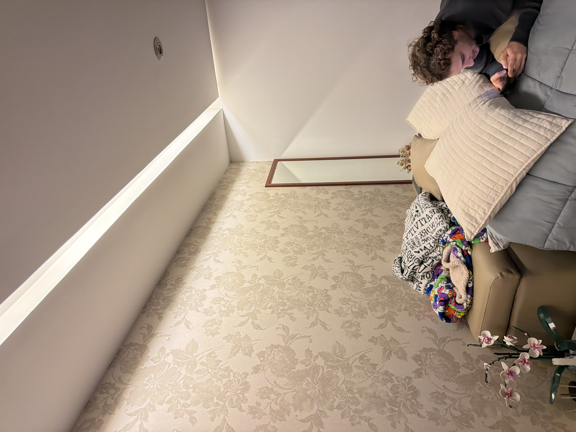 The scene depicts an indoor room in San Bernardino Tlaxcalancingo, Mexico. The ceiling is white, featuring a long recessed strip light along one edge and a circular recessed spotlight further inward. The left wall is covered in a beige floral wallpaper with a repeating pattern of large flowers and leaves. The adjacent wall is plain white.
In the foreground, a young adult male with medium-length curly brown hair, wearing a dark long-sleeved shirt, is lying prone on a bed. He is propped up by two quilted beige pillows and a plain beige pillow. He holds a smartphone with both hands and is looking down at its screen. The bed is covered with a light blue or grey comforter.
To the left, a tan or light brown ottoman is stacked with a white blanket patterned with black text, a colorful patterned blanket beneath it, and a single beige fluffy slipper. A tall, narrow mirror with a dark wooden frame leans against the floral wallpaper in the background. In the lower left, an artificial orchid plant with pink blossoms and green stems is displayed in a dark-colored vase on a small, dark surface.