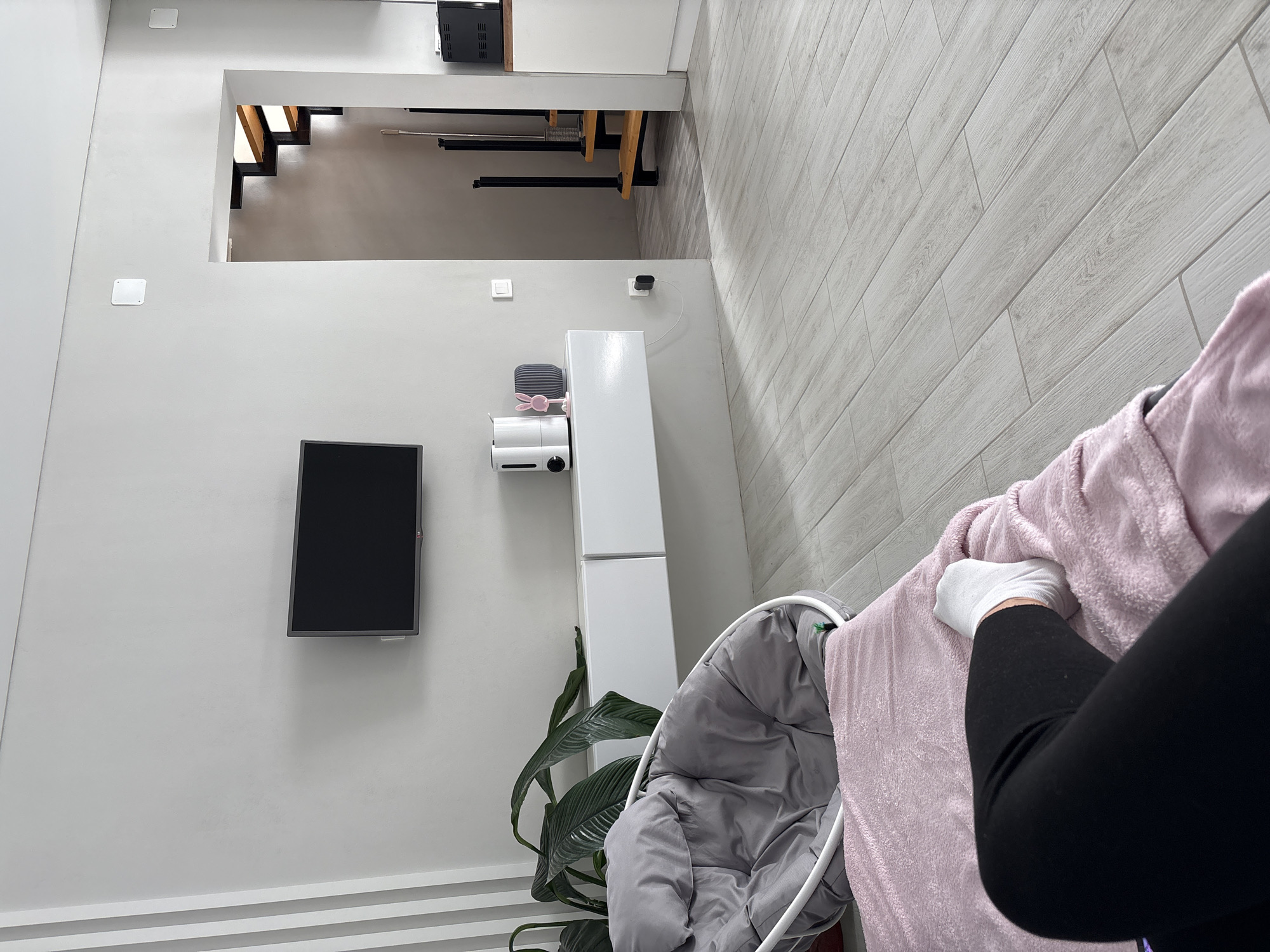 The image captures a modern residential interior in Votkinsk, Russia. The scene is viewed from a low perspective, focusing on a living area with an adjacent kitchen and stairwell. In the immediate foreground, the lower legs of an individual, clad in black leggings and a white sock on the visible foot, rest on a pink, plush blanket.
The floor throughout the visible space is surfaced with light grey, wood-look plank tiles. A white-framed, grey papasan-style chair with matching cushions is positioned in the mid-ground to the left. Behind the chair, a large green houseplant with broad leaves is visible. Mounted centrally on a light grey wall is a black flat-screen television. Directly beneath the television, a white floating media console with two compartments is affixed to the wall. On the console, from left to right, are a white and black appliance, a pink rabbit-shaped figurine, and a dark grey ribbed vase. A white light switch is located on the wall to the right of the console, with a black power adapter plugged into an outlet beneath it.
To the right, an open doorway leads to a stairwell featuring light wooden treads and black metal supports. Adjacent to the doorway on the right, a section of a white kitchen cabinet and a black appliance are visible, suggesting an open-plan kitchen area. The walls are uniformly painted a light grey.