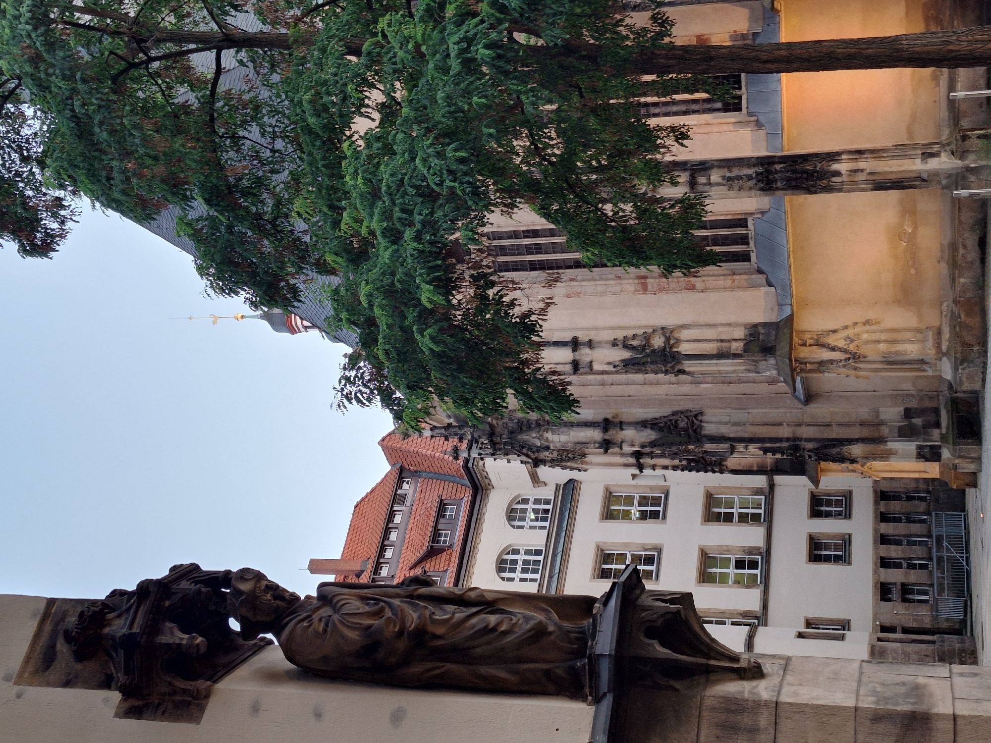 This is a vertical, eye-level, medium shot of a statue in front of a building and trees. The statue is made of dark bronze and depicts a cloaked figure seated on a pedestal with an ornate, gothic-style base. The building behind the statue is light-colored with several windows, some of which are arched. The roof of another building, with red tiles, is visible to the left. A large tree with lush green foliage extends into the frame from the top left, partially obscuring the buildings. The sky is overcast and pale. The overall impression is one of historical architecture and art in an urban setting. The lighting suggests it is late afternoon or early evening. No text is visible in the image. The location is identified as Chemnitz, Germany.
