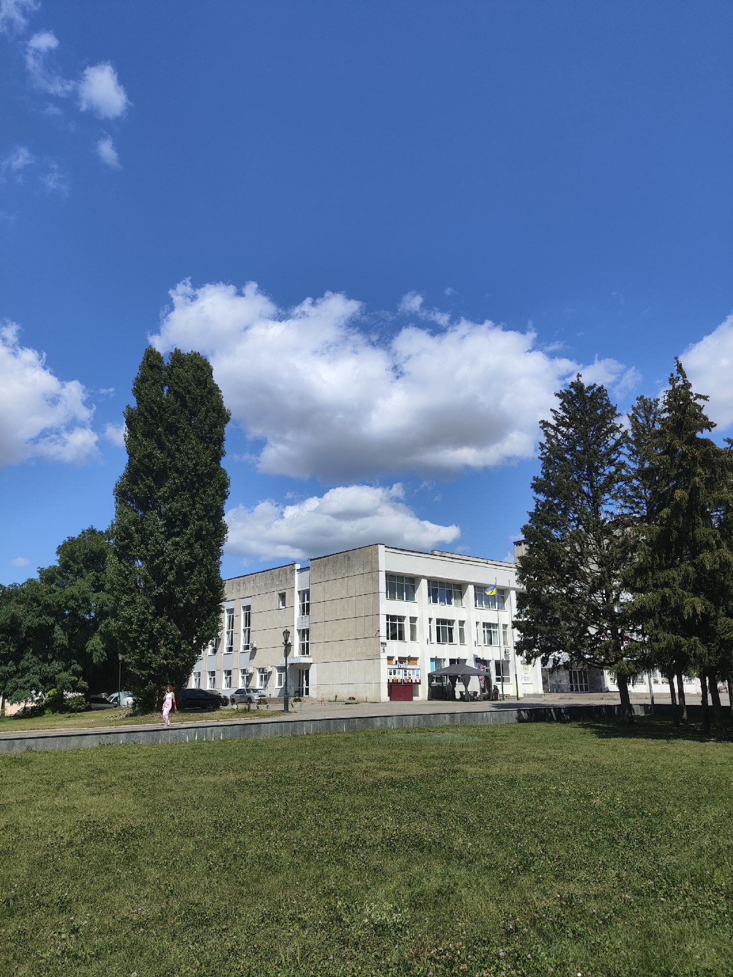 The image depicts a wide-angle, sunlit exterior view of a modern, light-colored, multi-story building situated in an urban environment. The building's facade is primarily concrete with numerous rectangular windows and several visible air conditioning units. A Ukrainian flag is visible flying from a pole on the building. Adjacent to the building are several mature evergreen trees and a very tall, slender deciduous tree. A grassy area with some sparse flowers is in the foreground, separated by a low wall and sidewalk from the building and trees. A person, appearing to be a woman in light-colored pants and a top, is walking on the sidewalk in front of the building. Several cars are parked in front of the building, and a dark-colored gazebo-like structure is also present. The sky is blue with scattered white cumulus clouds. The scene is consistent with the area around the Kharkiv National Medical University, specifically near the main entrance of the pedagogical faculty building at Prospekt Nauky 5.